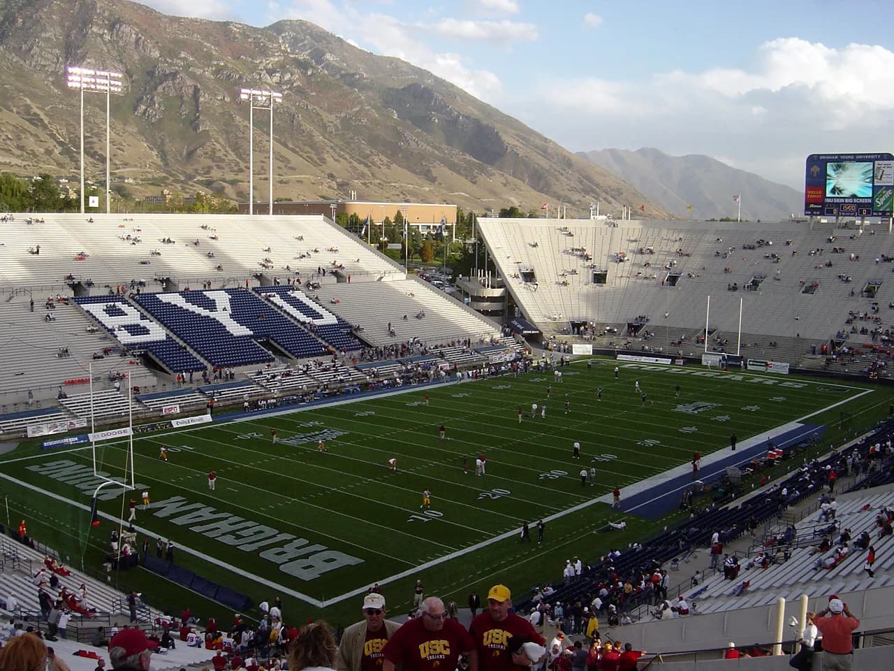 Wide view of a large college football stadium with BYU painted in the end zone, the letters BYU in the seats and mountains behind.
