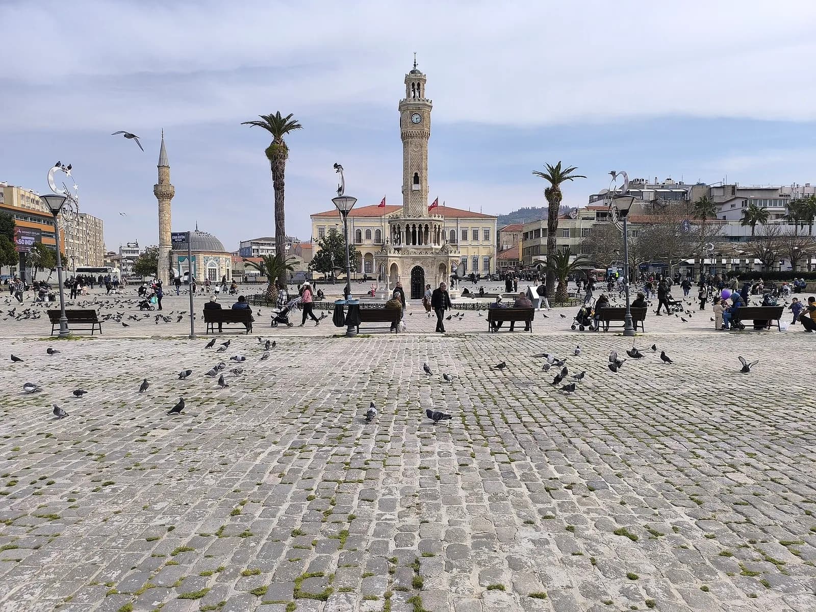 People sit near İzmir’s Clock Tower in Konak Square, with birds in the foreground.