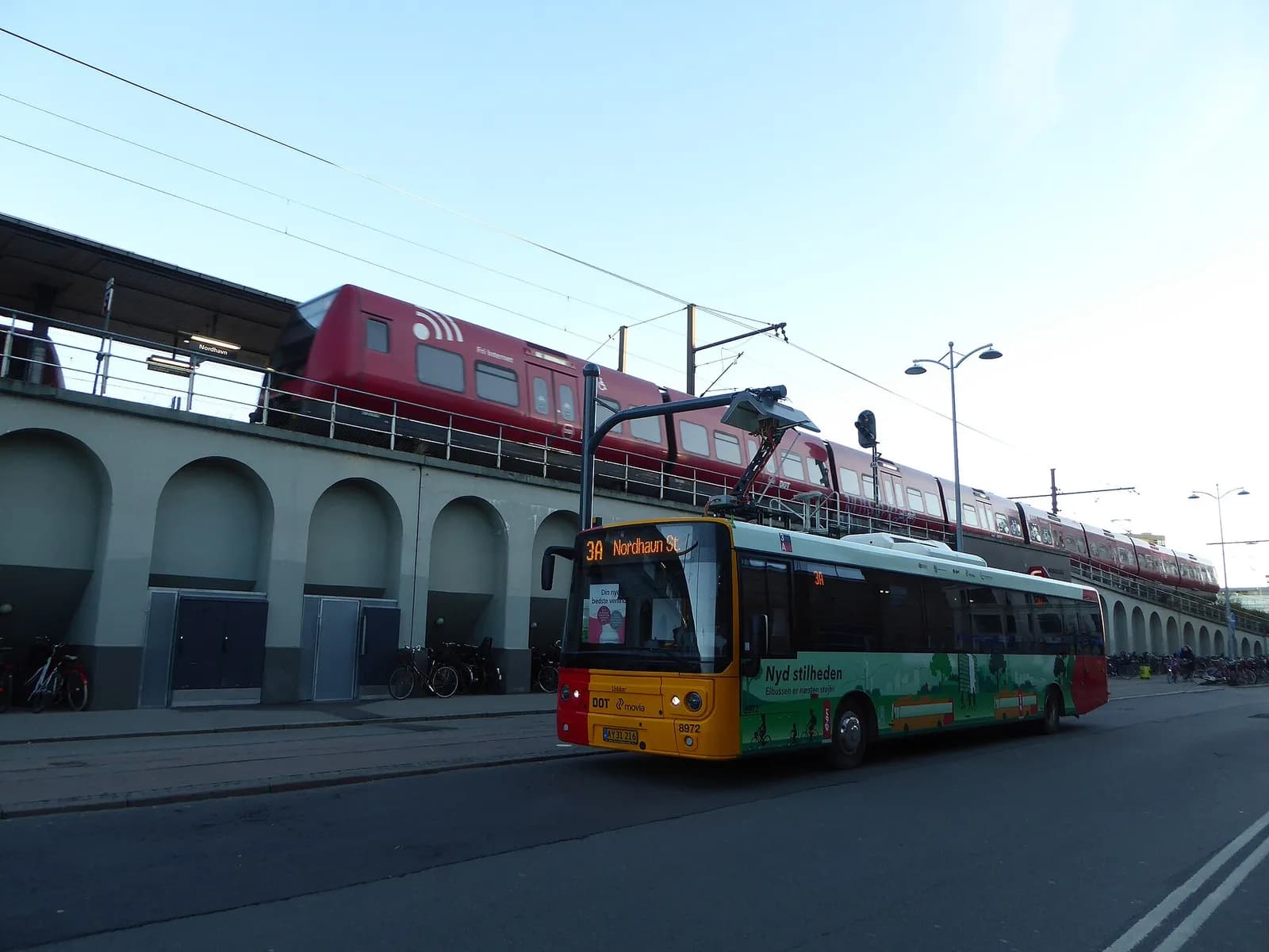 A Movia electric bus on route 3A at Nordhavn Station in Copenhagen, with an S-train passing above it.