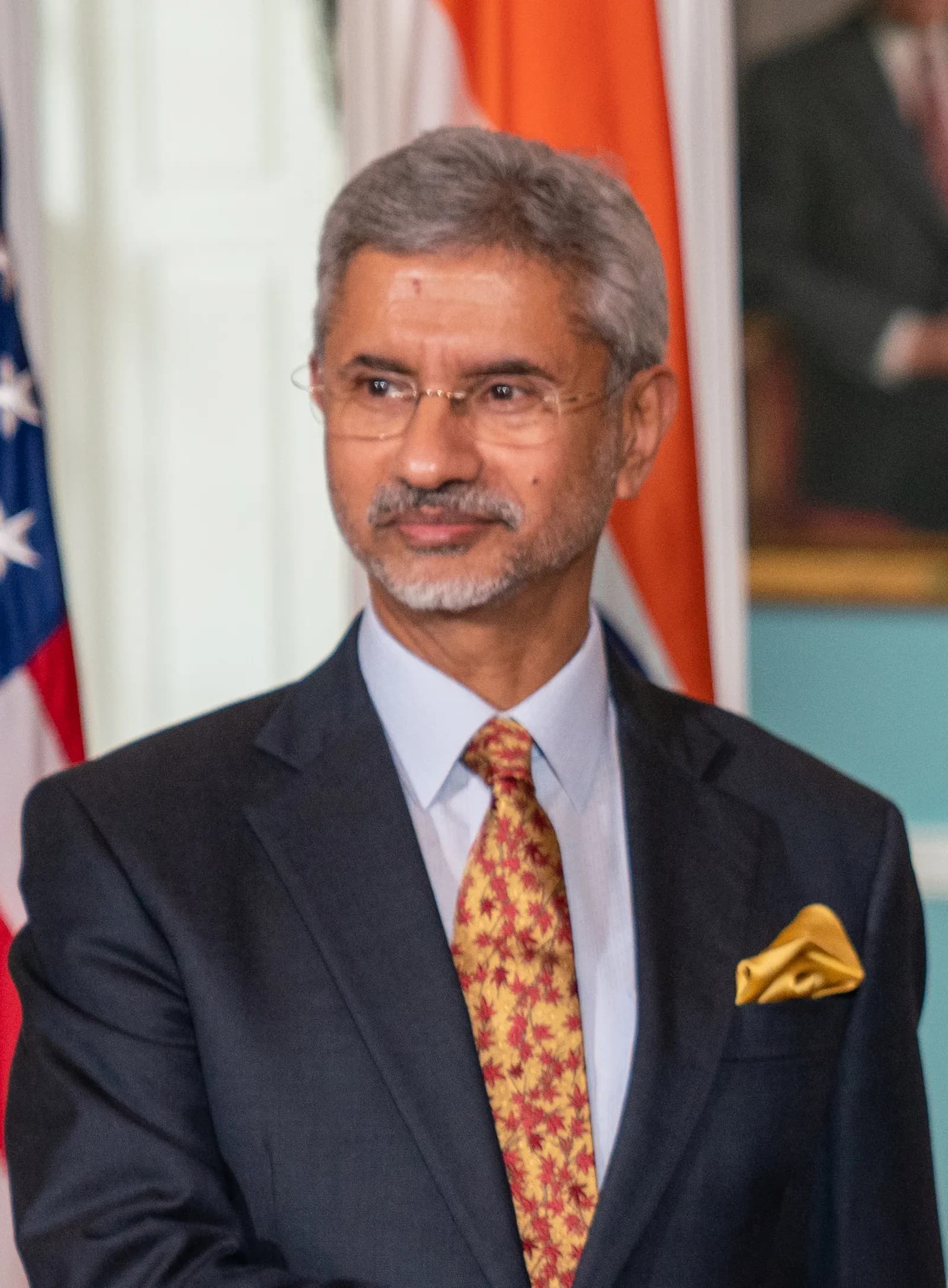 Close-up portrait of grey-haired Indian minister S. Jaishankar, wearing glasses and a dark suit with a patterned tie, standing in front of US and Indian flags.