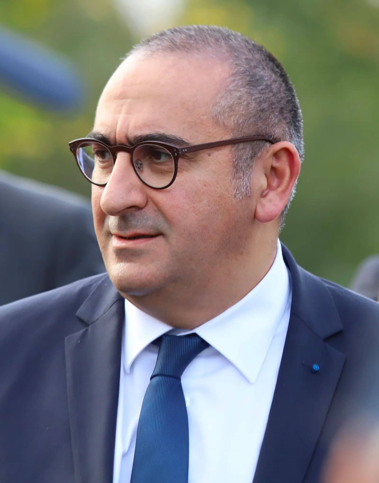 Close-up portrait of a balding man with dark-rimmed glasses, wearing a dark blue suit, white shirt and blue tie, looking to his left against a blurred green background.