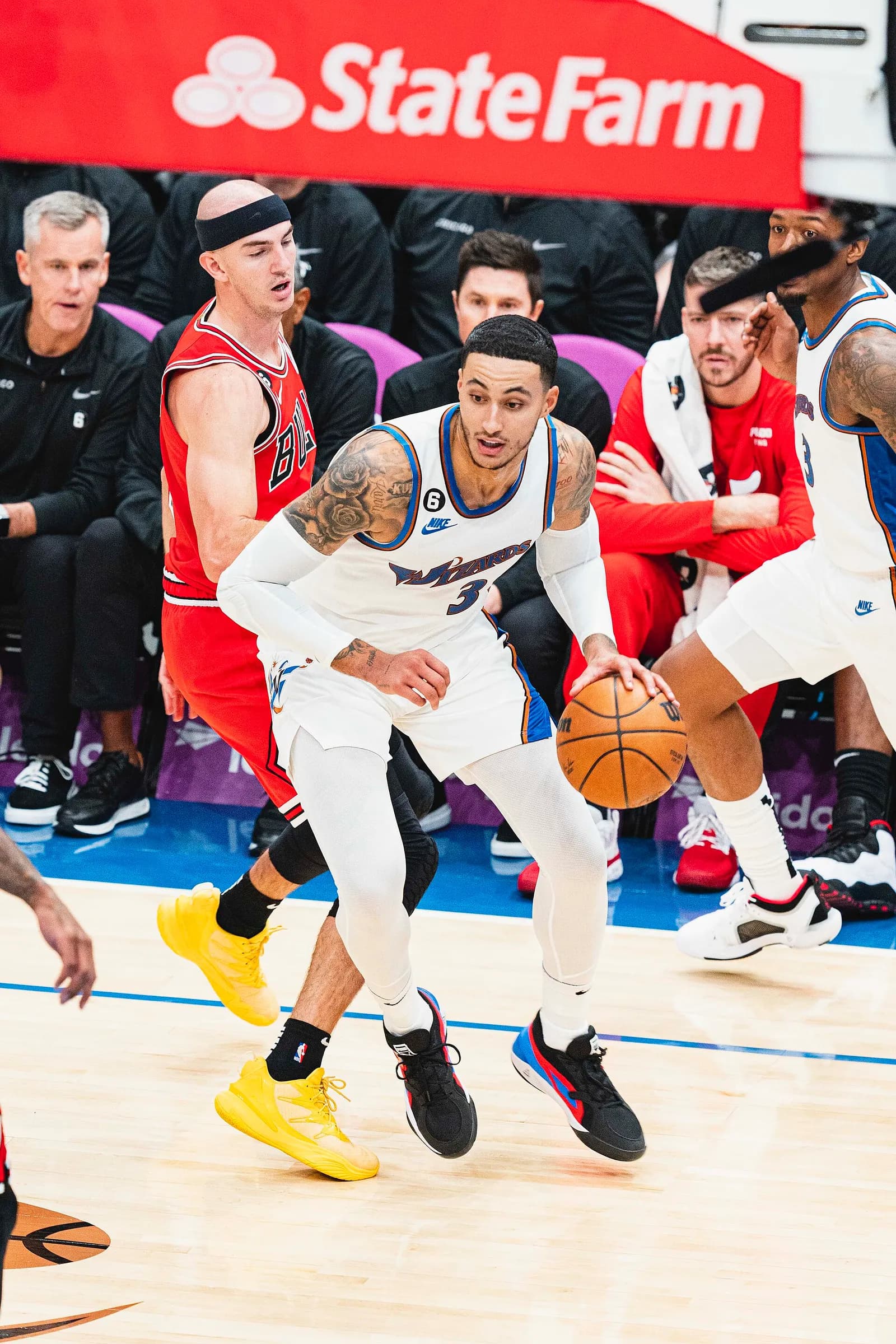 Basketball player in a red Bulls jersey with a headband defending a tattooed Wizards player in a white jersey who is dribbling the ball, with spectators in the background.
