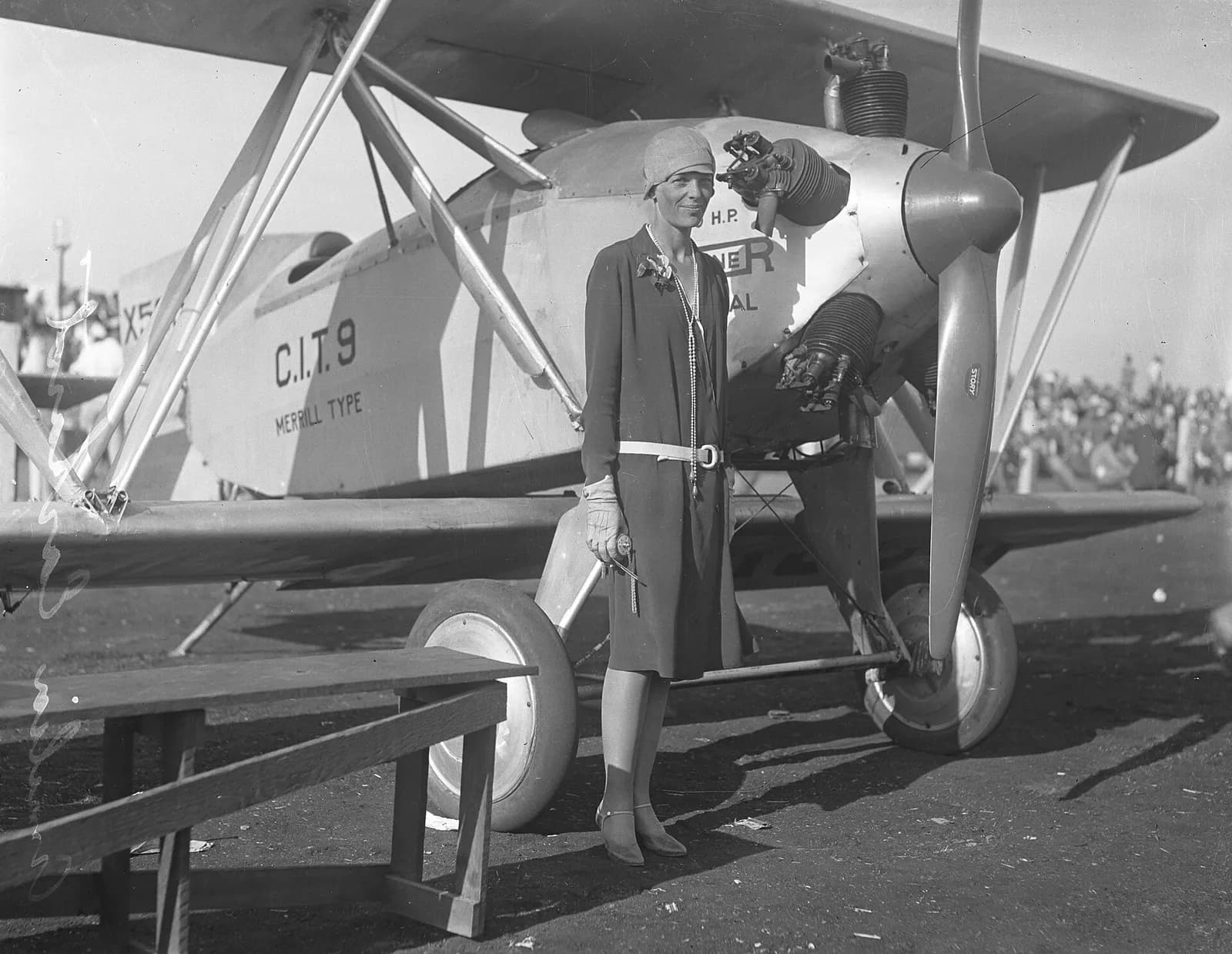 Black-and-white photograph of Amelia Earhart in a dark dress and close-fitting hat standing in front of a Merrill-type biplane marked C.I.T.9, with a wooden bench in the foreground.
