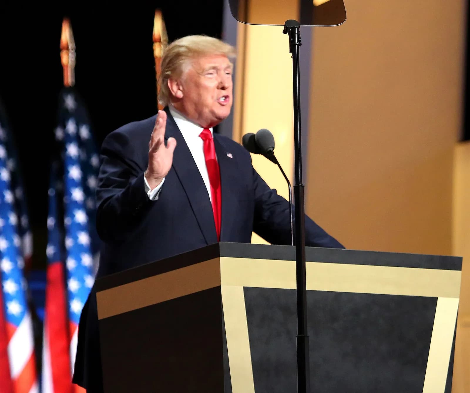 Donald Trump, wearing a dark suit and red tie, speaks at a dark podium with a raised hand, with an American flag behind him.