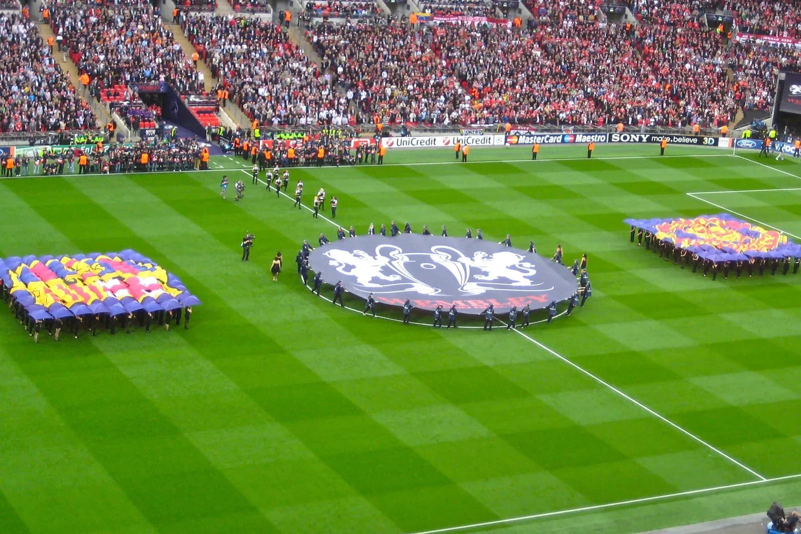 A large Champions League banner is displayed on the Wembley pitch before the 2011 final between Barcelona and Manchester United, with club-coloured banners on either side and a full stadium crowd.