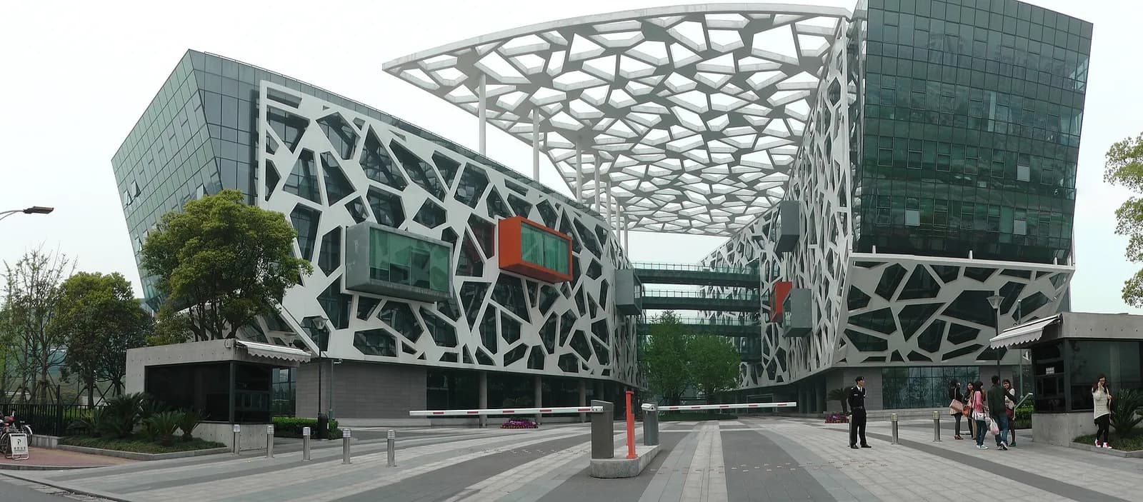 Modern glass office buildings with white lattice-patterned cladding and a large lattice canopy over an entrance plaza, with a few pedestrians in the foreground.