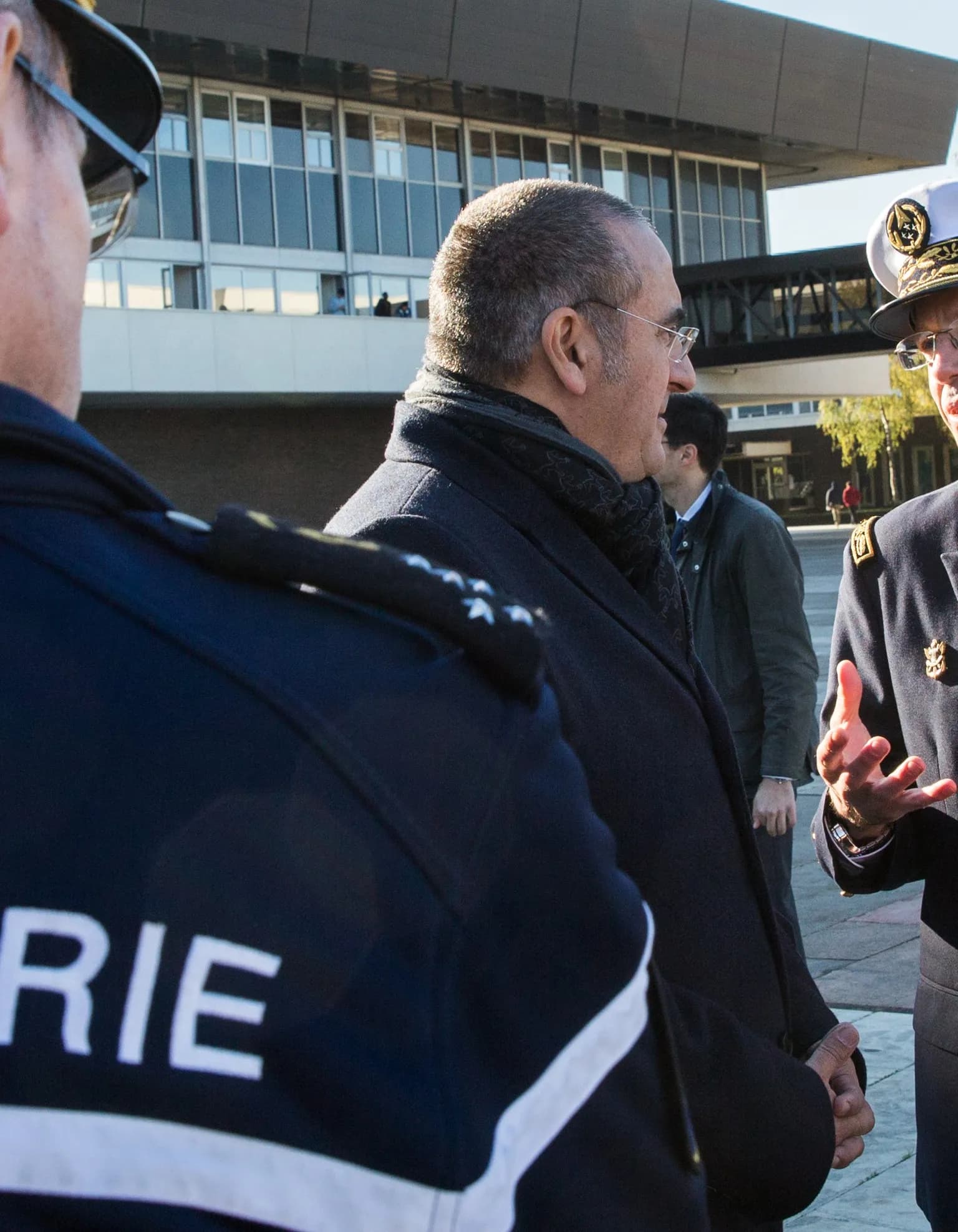 A man in a dark coat and patterned scarf, wearing glasses, stands in profile in a sunlit outdoor courtyard between uniformed gendarmerie officers, with a glass-fronted building behind.