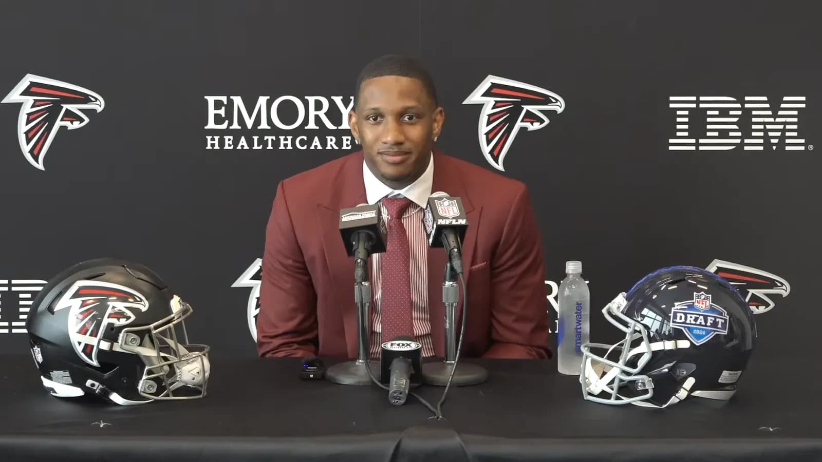 Quarterback Michael Penix Jr. in a maroon suit and tie seated behind microphones at a press conference table flanked by two Atlanta Falcons helmets.