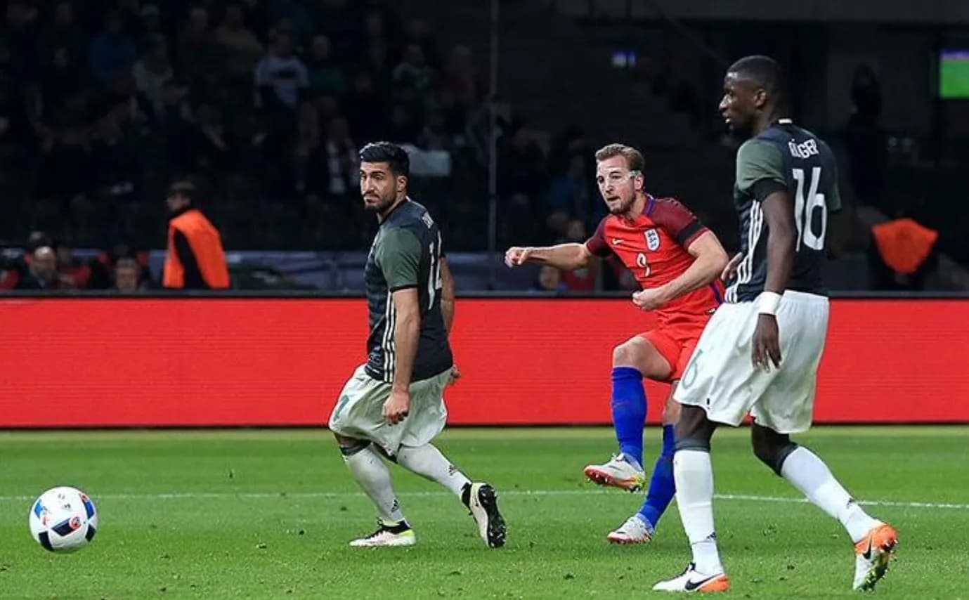 Harry Kane in a red England shirt and blue socks running between two Germany players in dark shirts and white shorts, with the ball on the grass to the left.