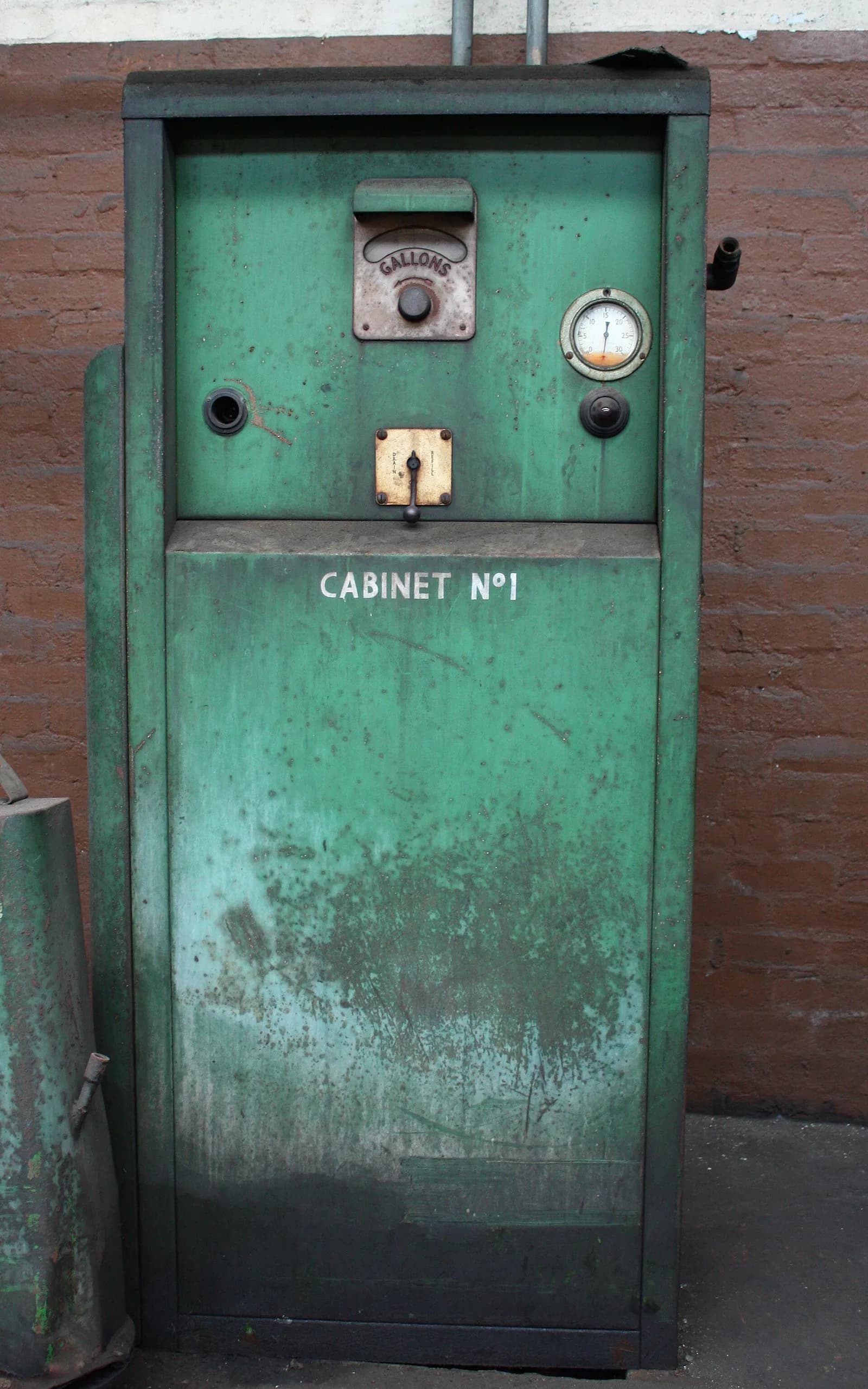 An old green diesel pump with a gallon indicator and gauge stands against a brick wall.