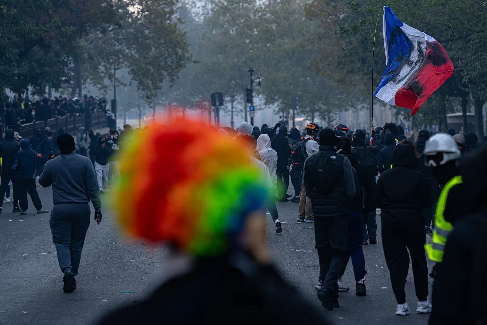 A protester wearing a rainbow clown wig walks among a crowd, with a French flag and tear gas in the background.
