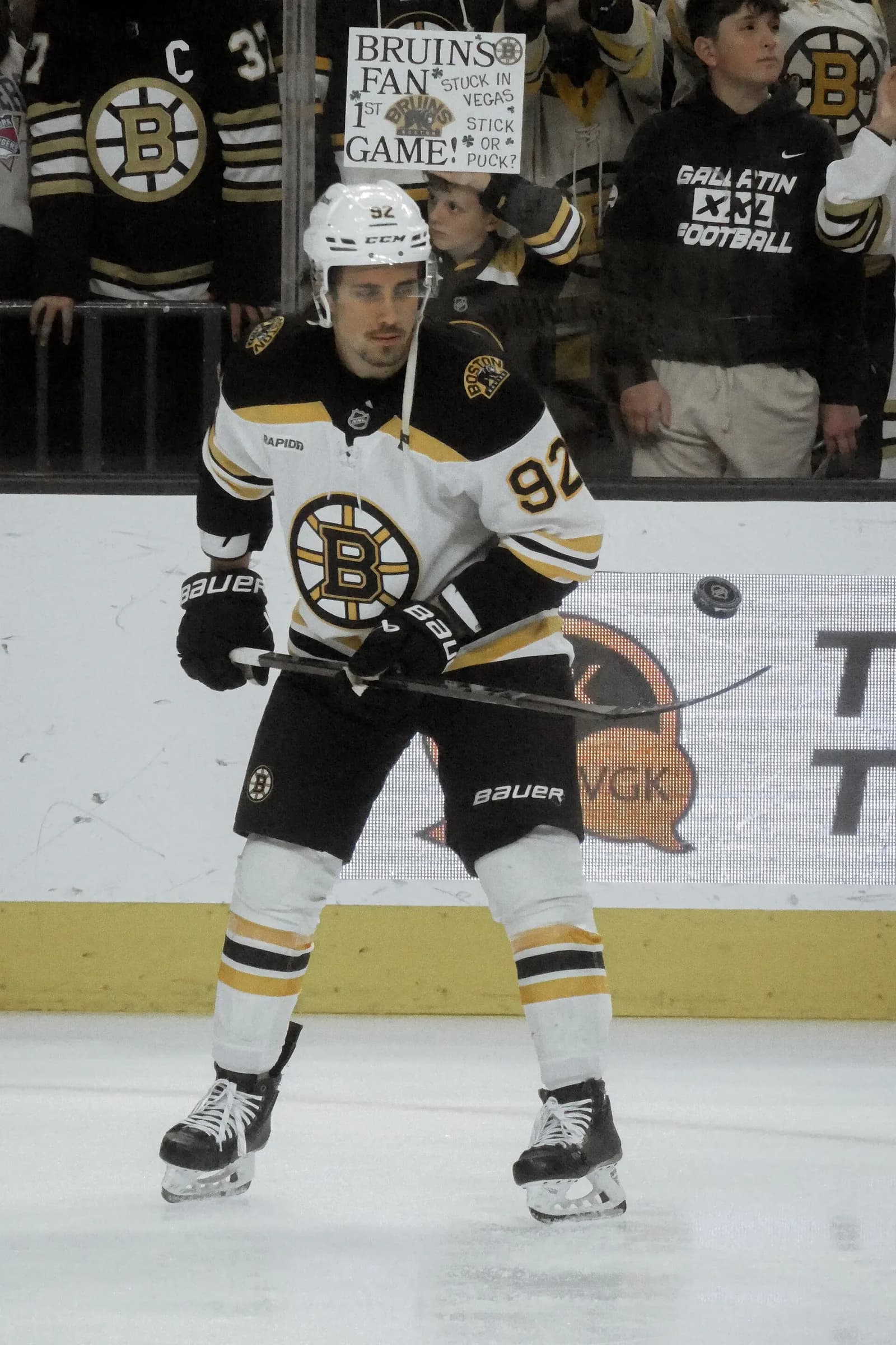 A hockey player in a white Boston Bruins jersey with black and gold trim, number 92, stands with his stick on the ice during warm-ups in front of spectators holding a sign behind the glass.