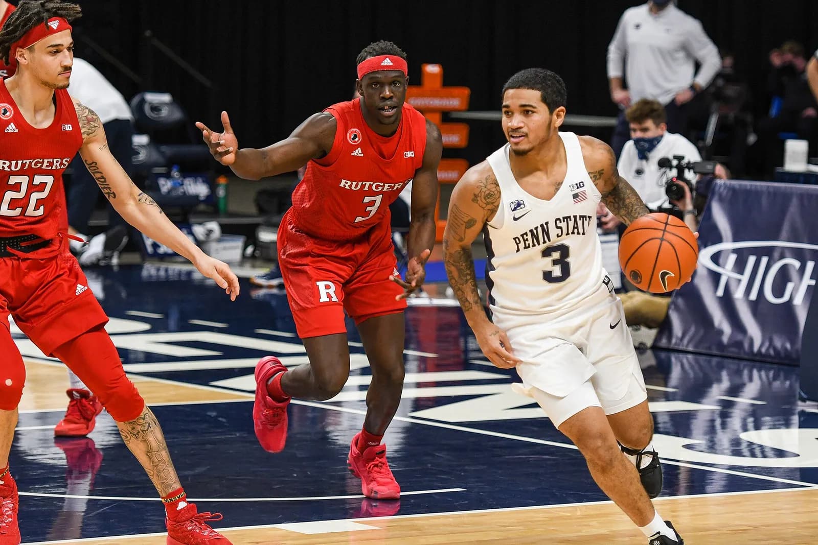 Two Rutgers basketball players in red uniforms defending a Penn State player in a white jersey who is dribbling the ball up the court.