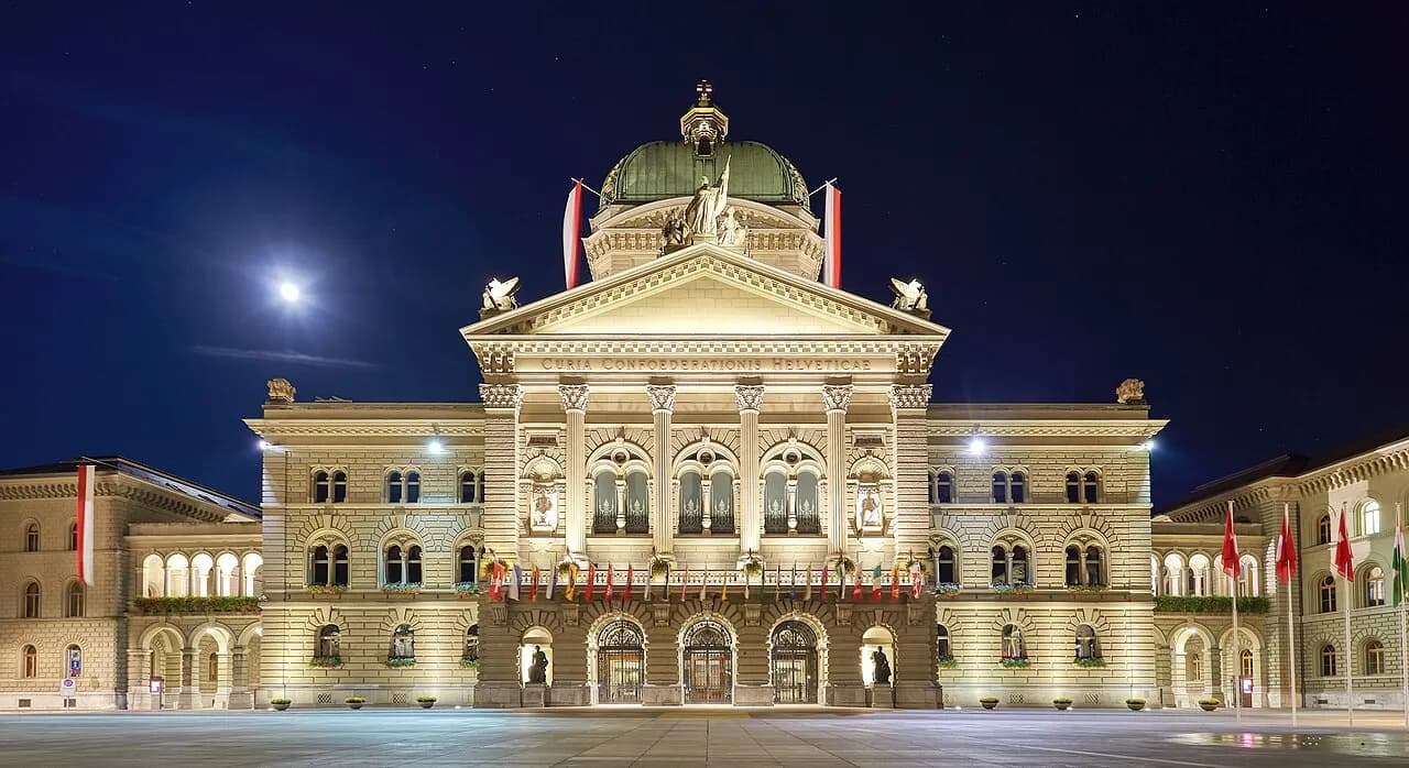 The illuminated Federal Palace of Switzerland and flags overlooking Parliament Square in Bern at night.