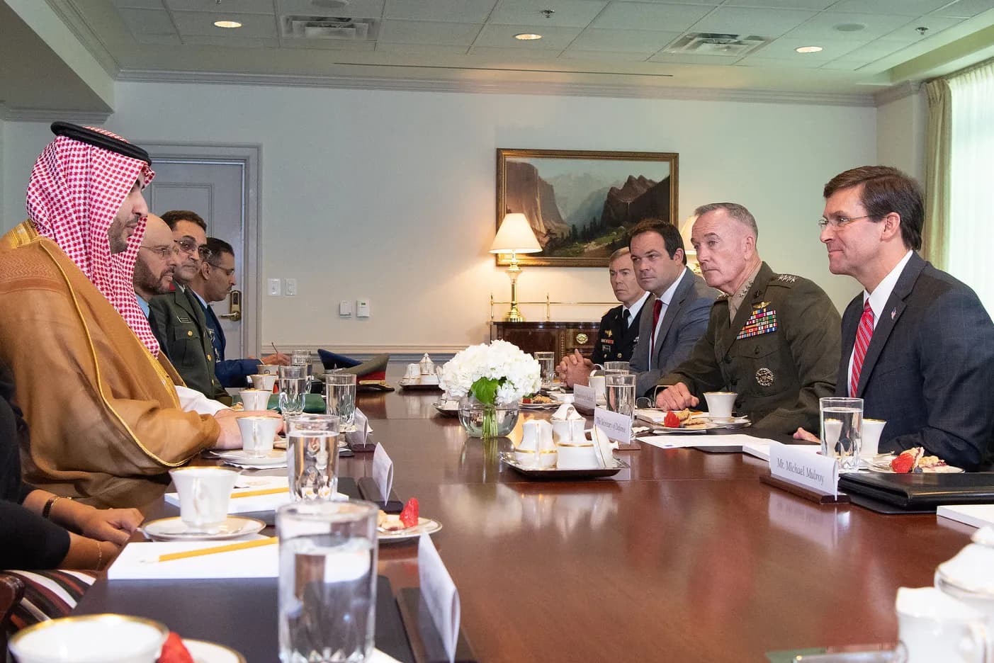 A man in a red-and-white checked headdress and beige robe sits at the left of a long polished conference table facing a group of men in suits and military uniform, including one general in olive green, in a room with a landscape painting on the wall.