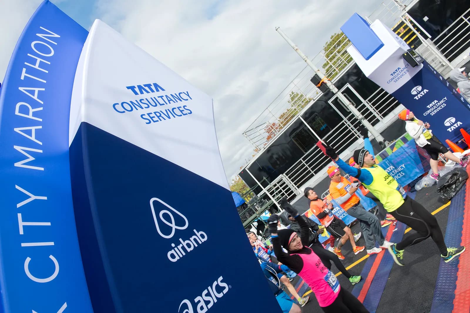 Runners in bright running kit, one with arms raised, standing near a large blue and white inflatable sign reading CITY MARATHON with sponsor logos at the start of the New York City Marathon.