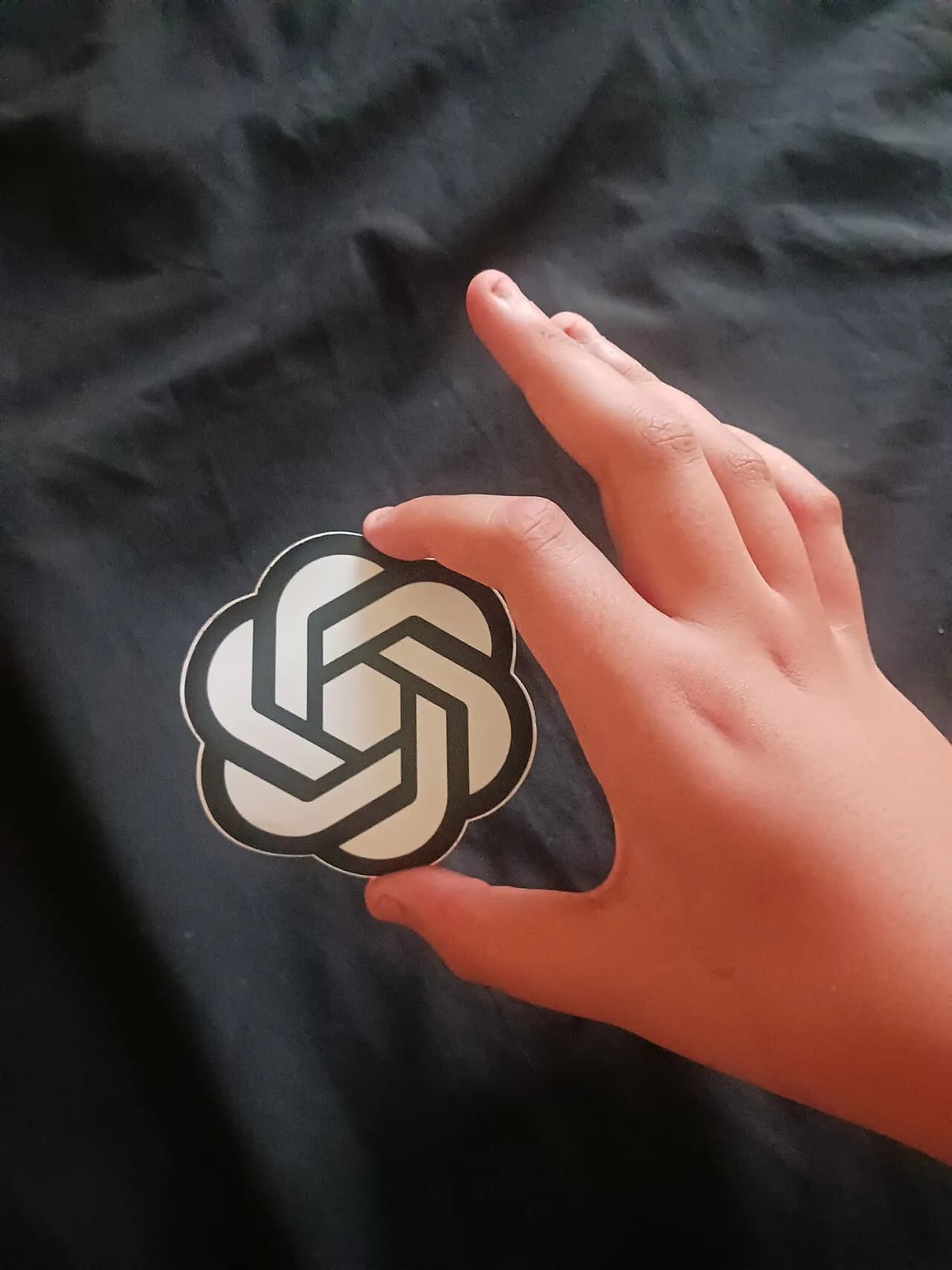 A hand holds a sticker bearing the OpenAI icon.