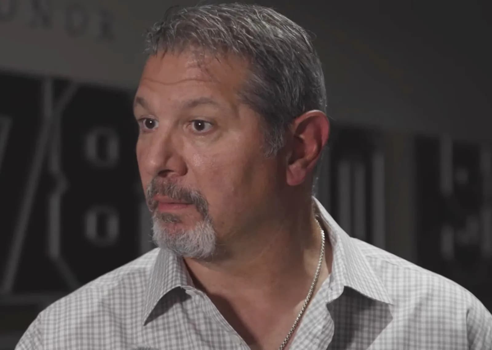 Close-up portrait of a grey-haired, grey-goateed man in a light checked shirt and silver chain, looking to the side during an interview.