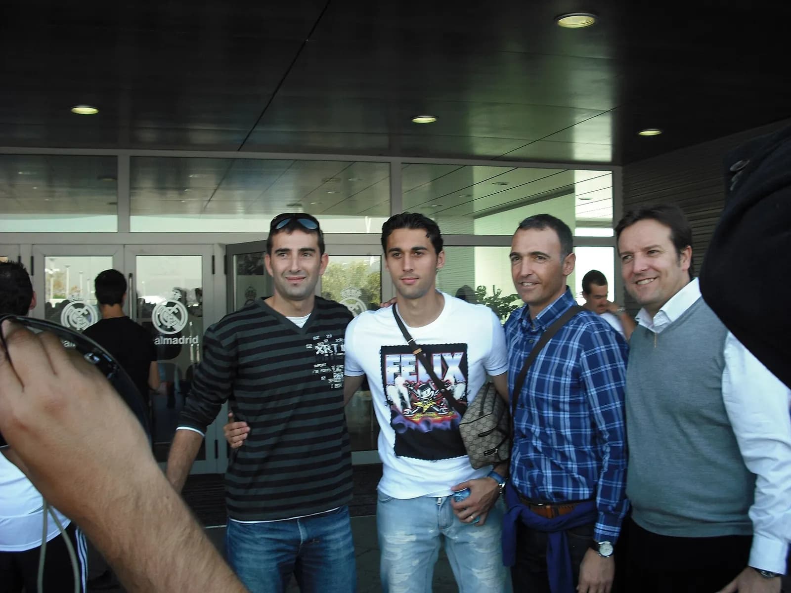 Four men standing side by side in casual clothes in front of glass entrance doors bearing a Real Madrid crest, with Arbeloa in a white printed T-shirt in the centre.