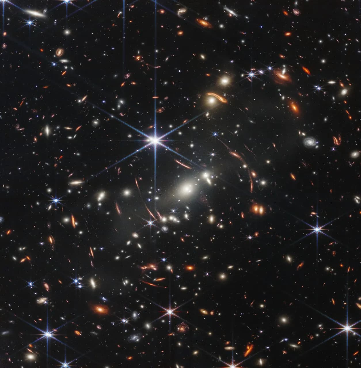 Webb deep-field image showing many distant galaxies, curved orange arcs and bright foreground stars