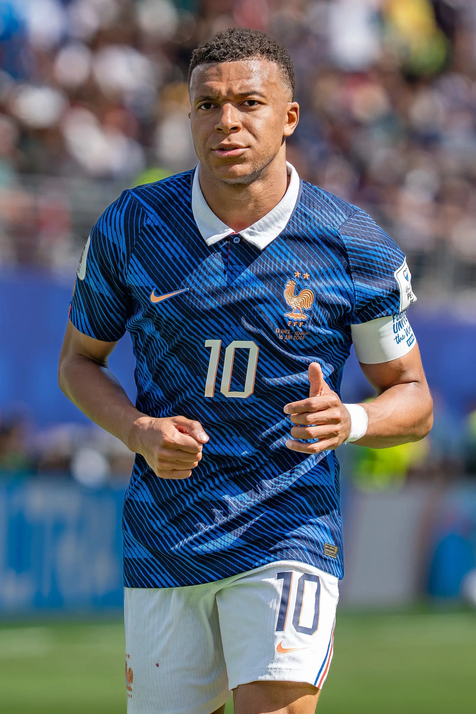 Footballer Kylian Mbappe, wearing a blue France number 10 shirt and white shorts, runs forward towards the camera with a blurred stadium crowd behind him.