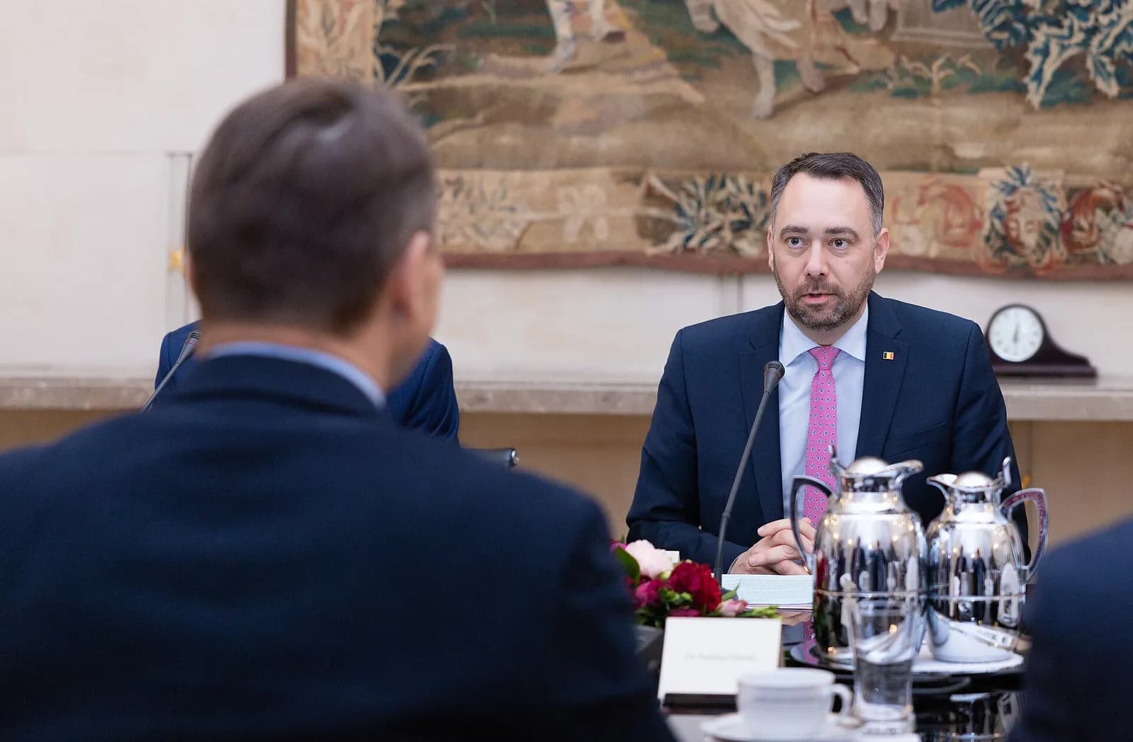 A bearded man in a dark suit and pink tie seated at a table with a microphone, facing another man seen from behind.