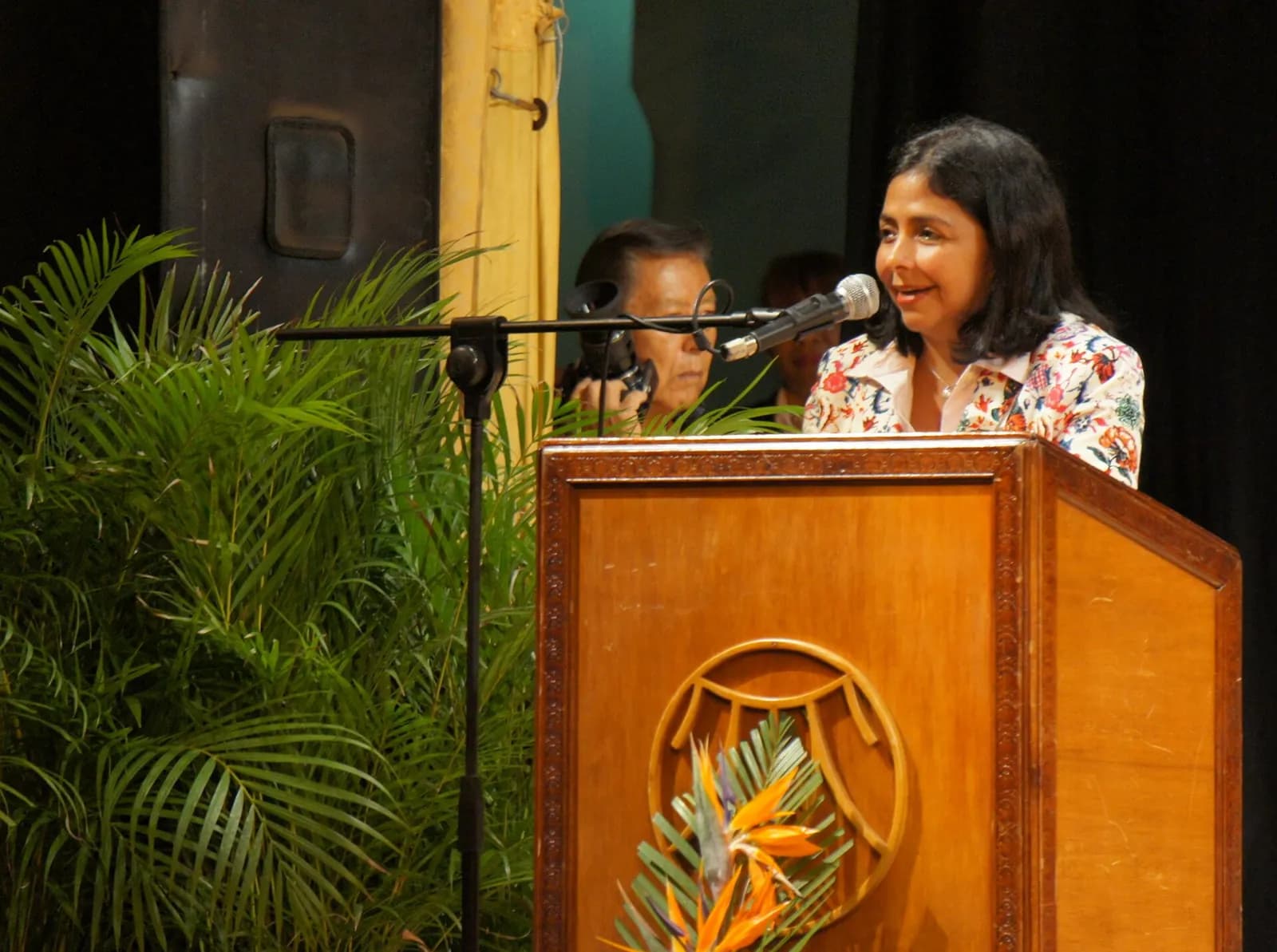 A woman with shoulder-length dark hair and a floral shirt speaks at a wooden lectern with a microphone, palm leaves to the left and a man behind her.