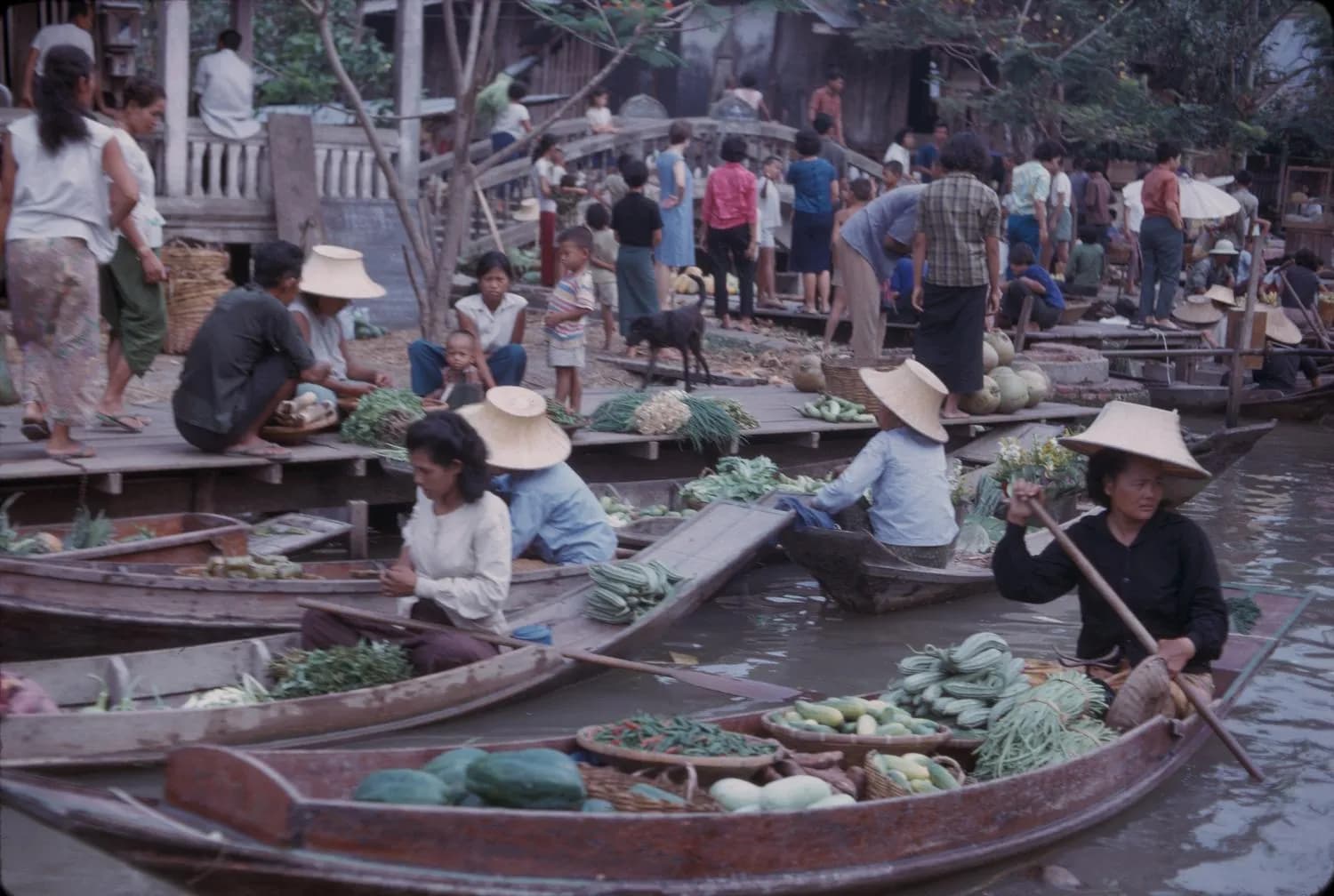 People sell fresh produce from boats at a floating market in Bangkok, with a crowd gathered along the dock.