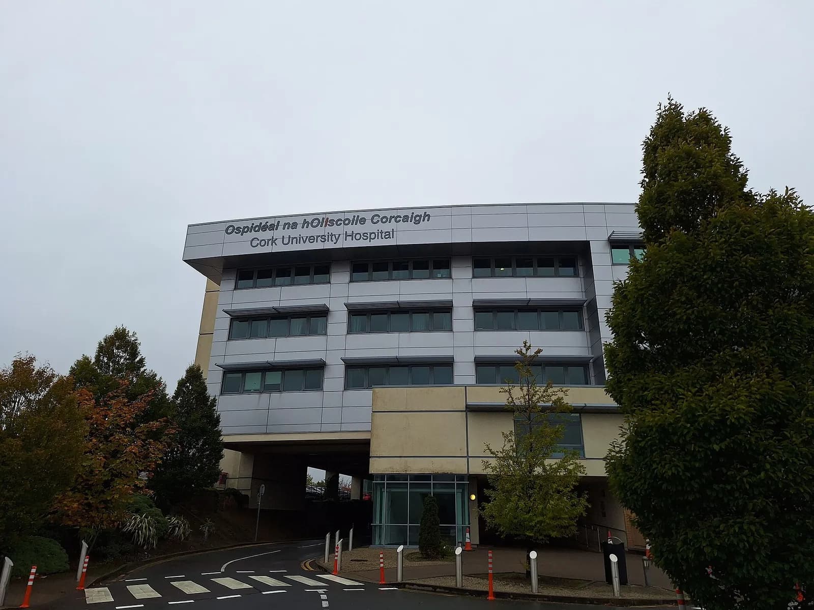 A multi-storey hospital building with grey panelling and rows of windows, with 'Ospidéal na hOllscoile Corcaigh Cork University Hospital' written on the top facade, above a road and trees.