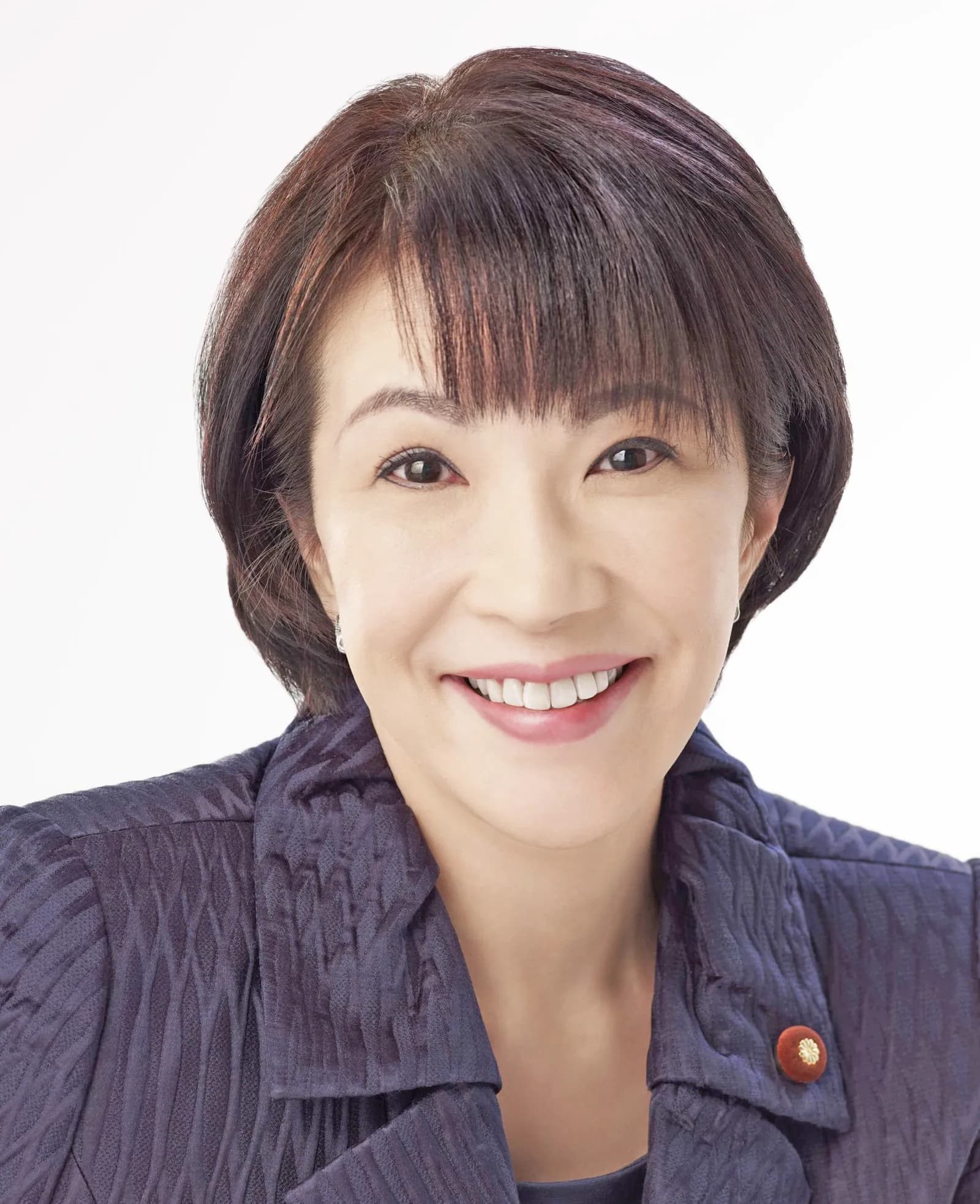 Head-and-shoulders portrait of Sanae Takaichi smiling against a white background, wearing a dark patterned jacket with a red pin on the lapel.