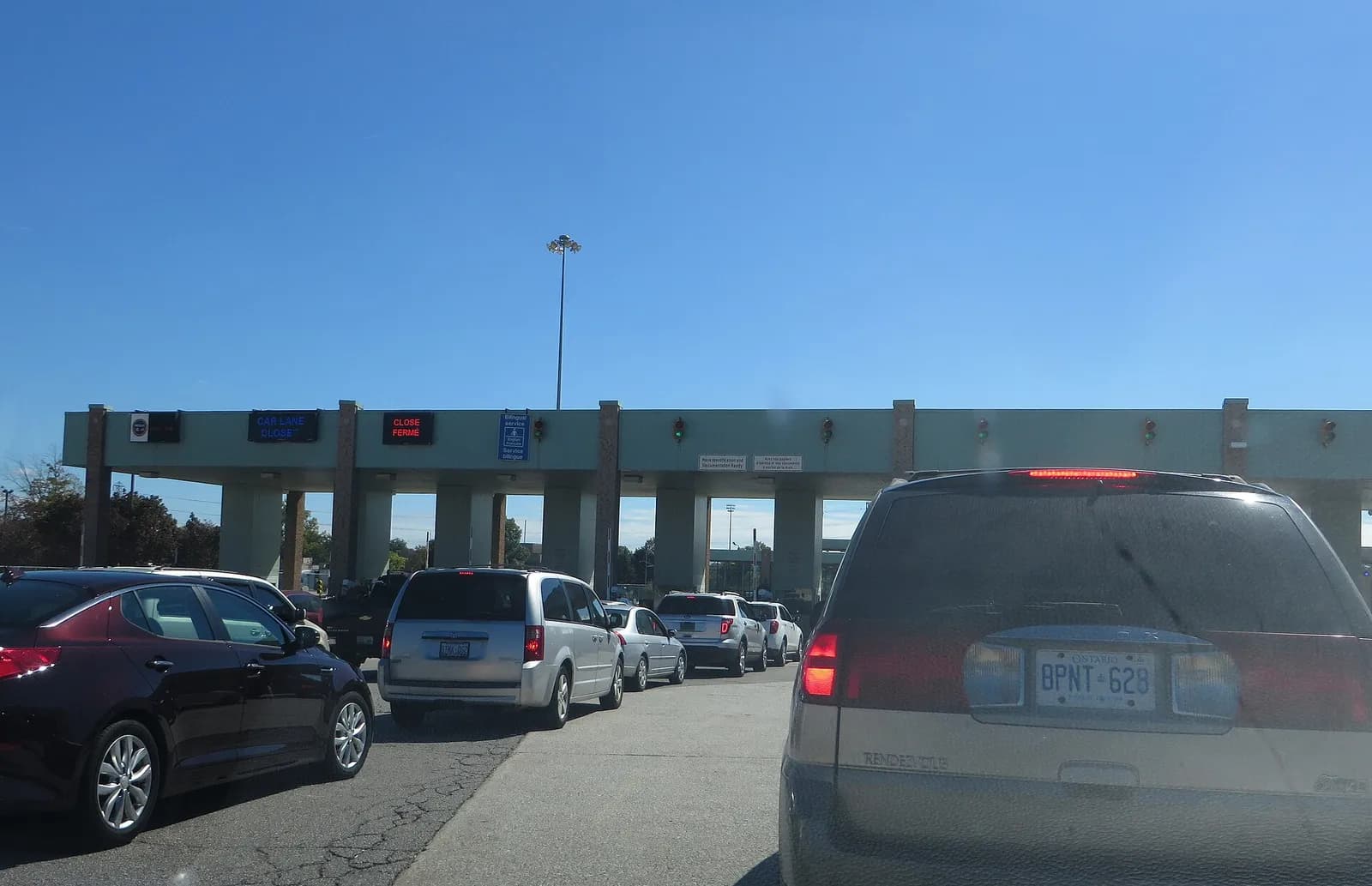 Cars lined up in several lanes at a Canada Border Services Agency toll-style inspection canopy, with red and blue lane signs above the booths and a clear blue sky.