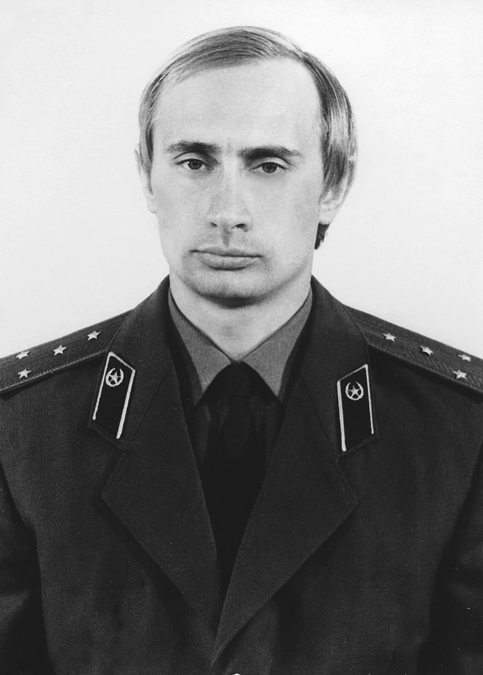 Black-and-white head-and-shoulders portrait of a young Vladimir Putin in a dark military uniform with KGB collar tabs and starred shoulder boards, looking straight at the camera.