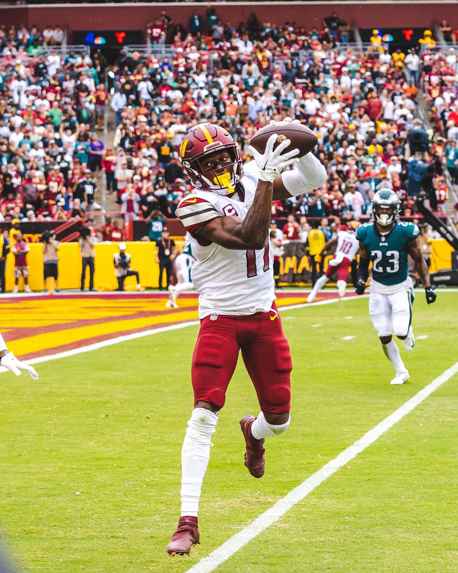 American football player in a burgundy and gold helmet and white jersey, number 1, leaping to catch the ball near the end zone with an Eagles defender running behind him.