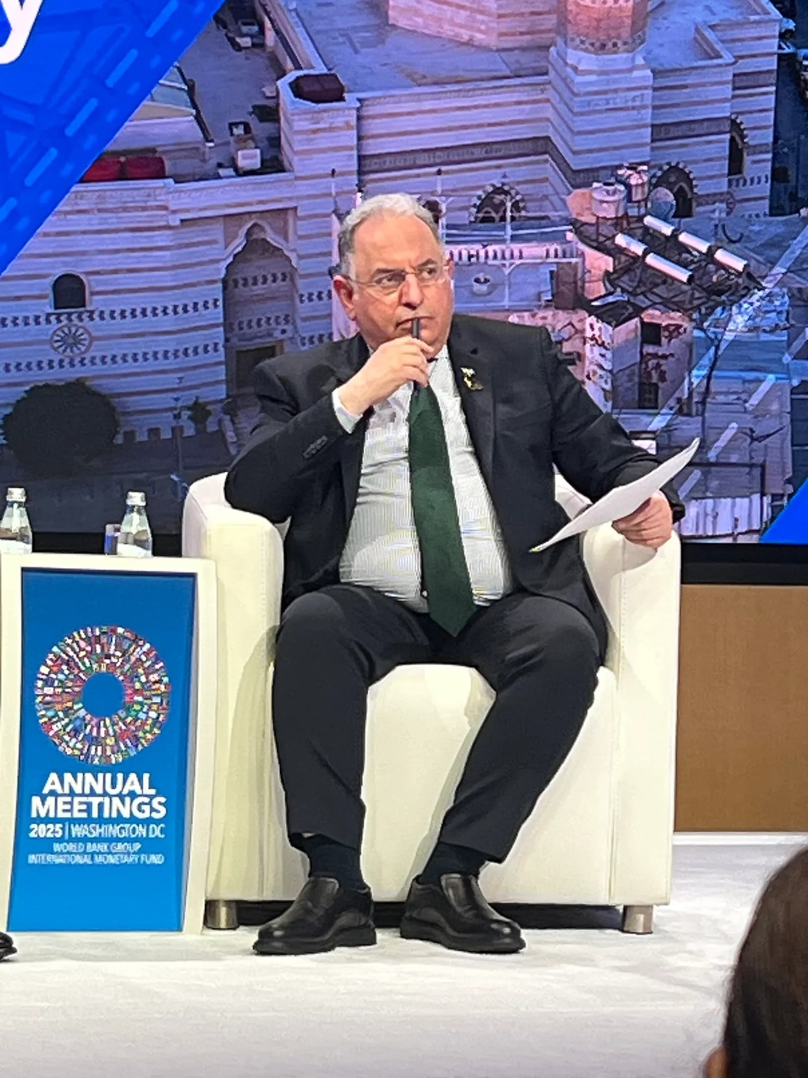 A man in a dark suit and green tie sits in a white armchair holding a microphone, beside a blue sign reading Annual Meetings 2025, Washington DC, World Bank Group and International Monetary Fund.