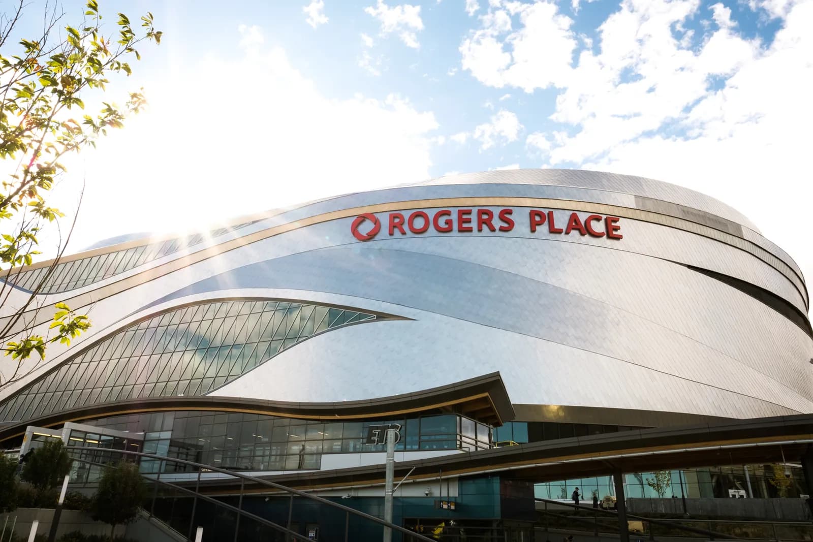 Curved silver metal exterior of Rogers Place arena with large red 'ROGERS PLACE' sign on the facade against a cloudy blue sky.