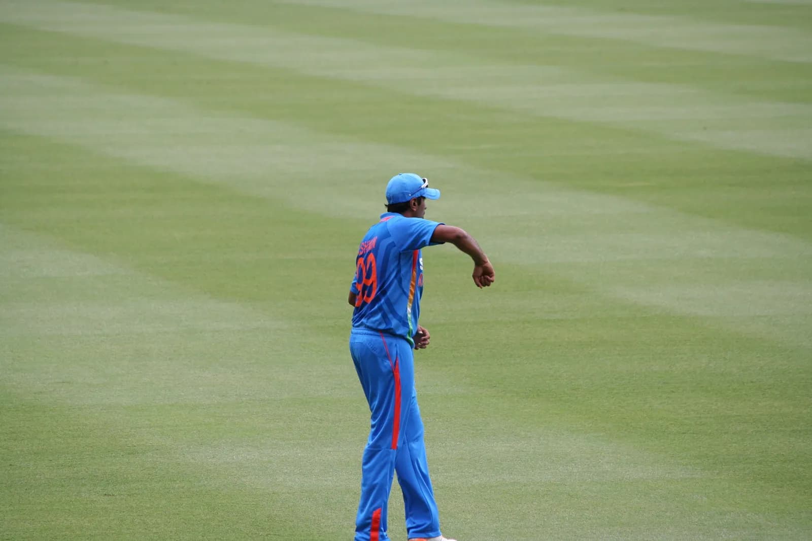A cricketer in a blue India shirt and cap, numbered 99 with ASHWIN on the back, stands on a green field facing away from the camera.