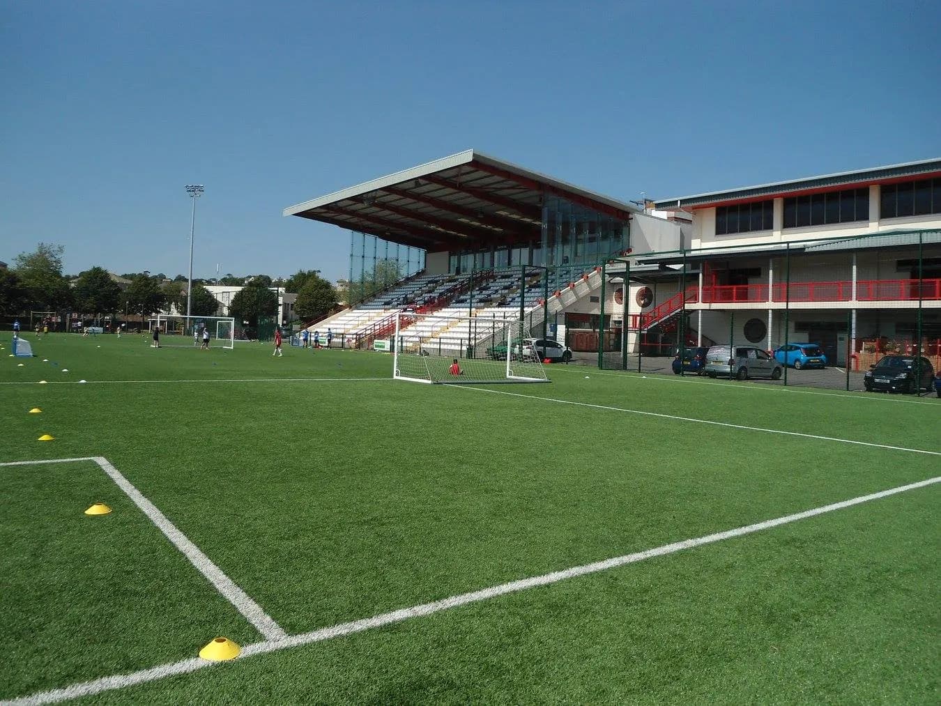 An empty artificial football pitch with yellow training cones in the foreground, a goal and a large covered main stand with seating terraces at Springfield Stadium under a clear blue sky.