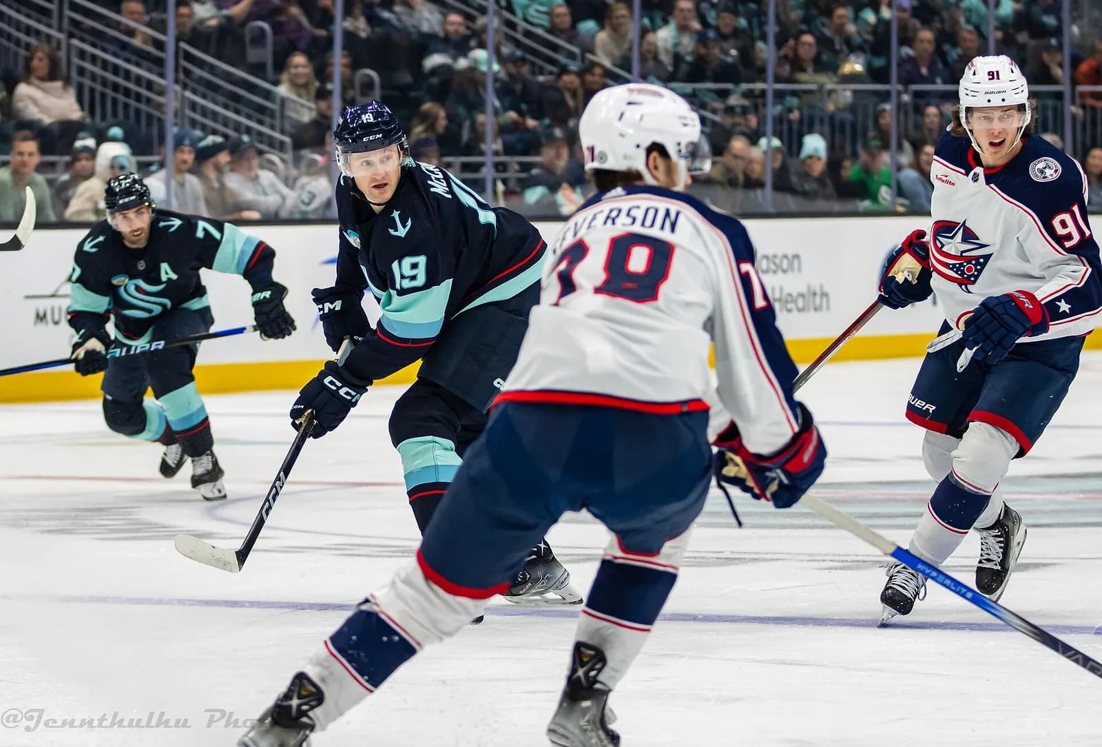 Ice hockey players in Blue Jackets white and Kraken dark blue jerseys during a game, with spectators in the background.