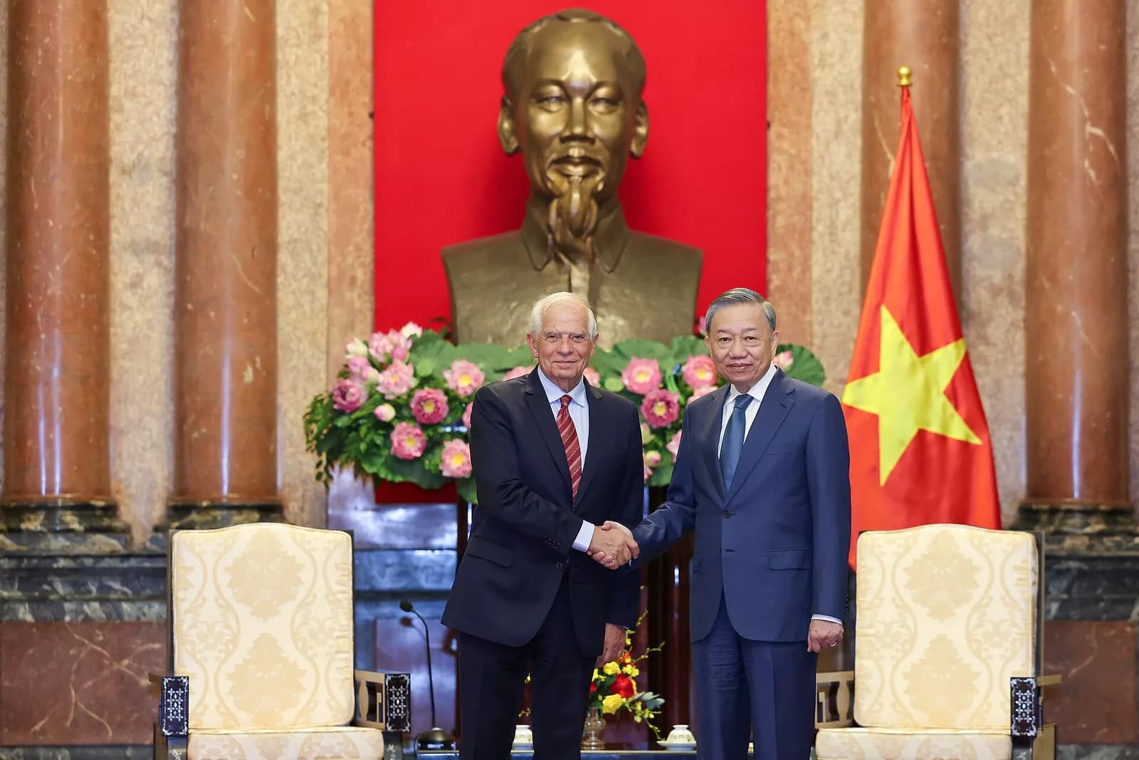 Two men in dark suits shake hands in front of a bust of Ho Chi Minh and a Vietnamese flag, between two ornate chairs.
