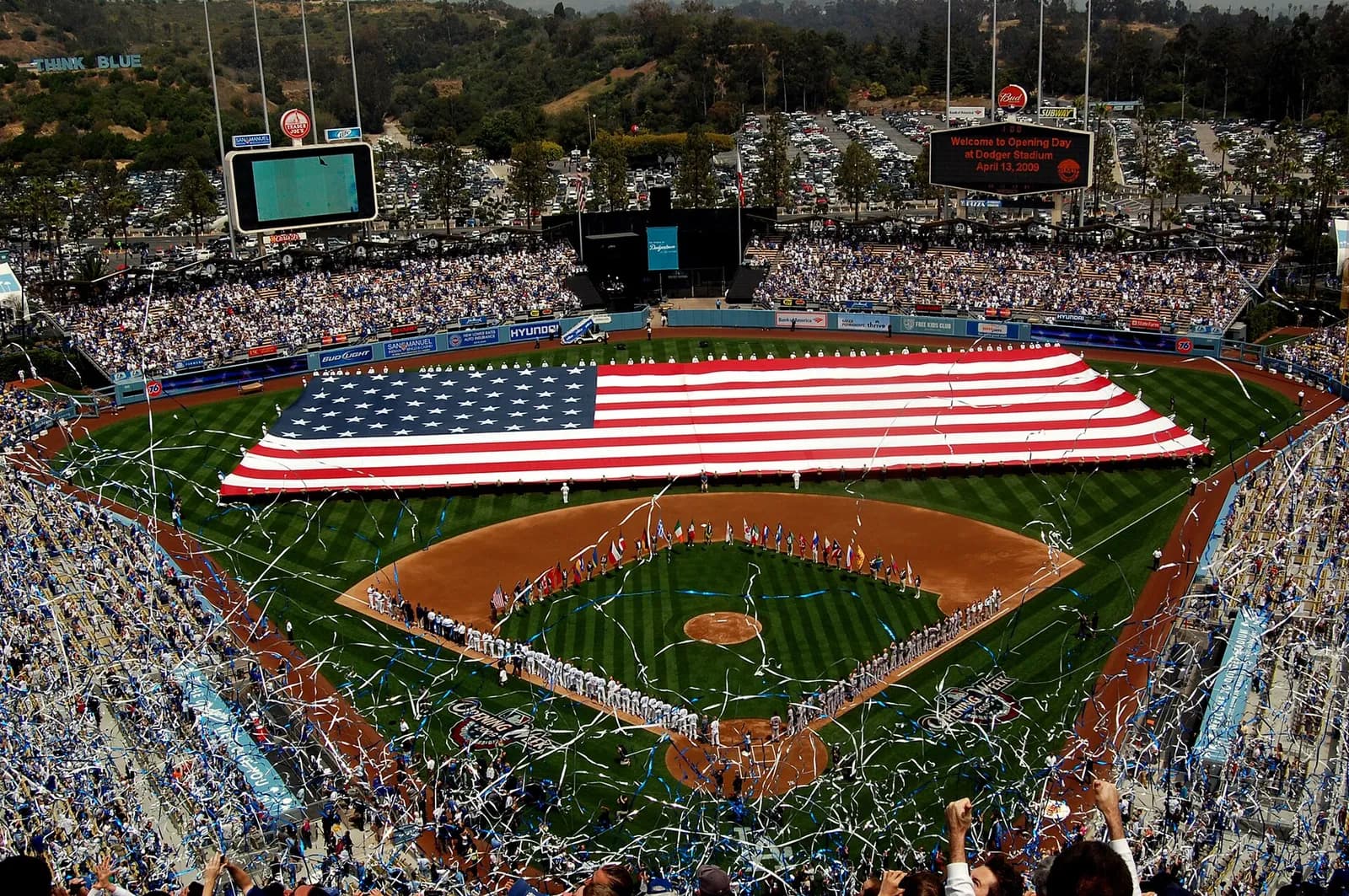 A large American flag is unfurled across the field at Dodger Stadium, with spectators in the stands.