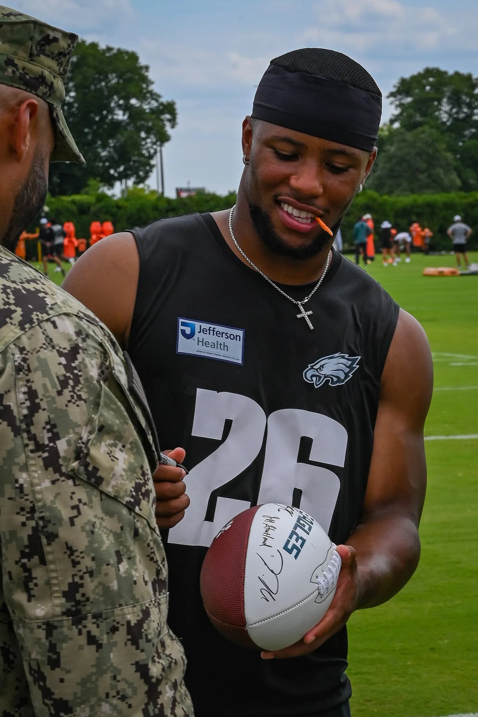 Saquon Barkley holds a signed football beside a person in uniform at Eagles training camp.