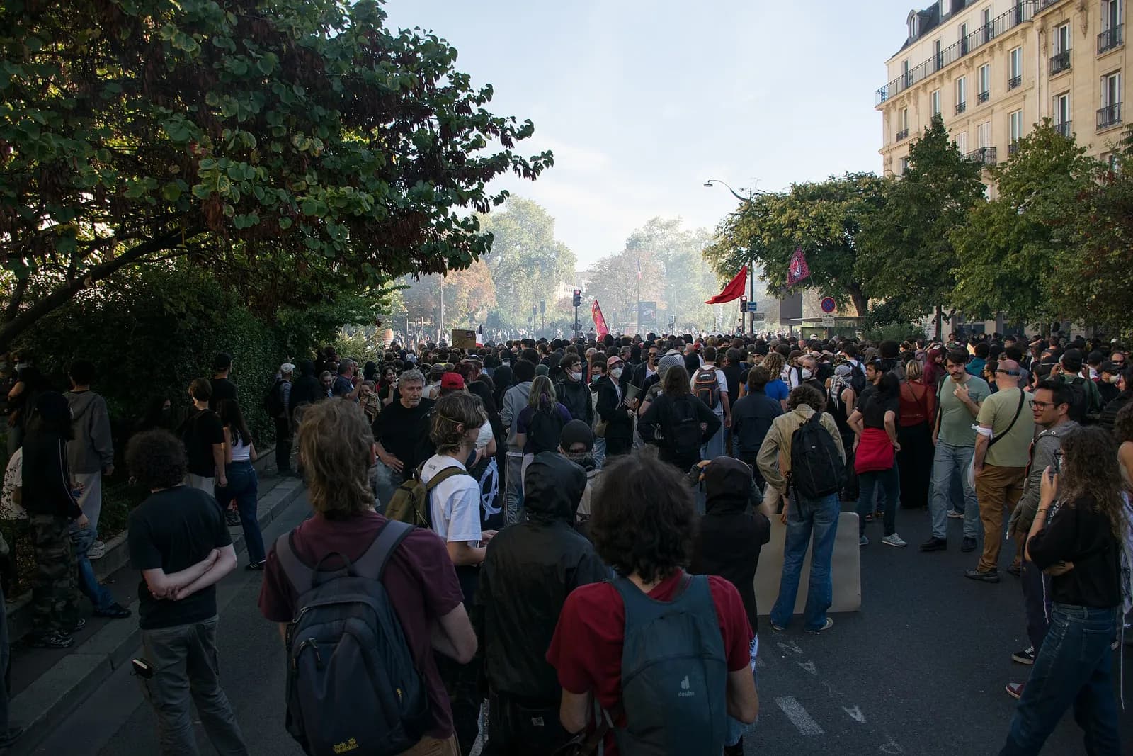 Students and other demonstrators gather along a tree-lined street in Paris, with red flags visible.