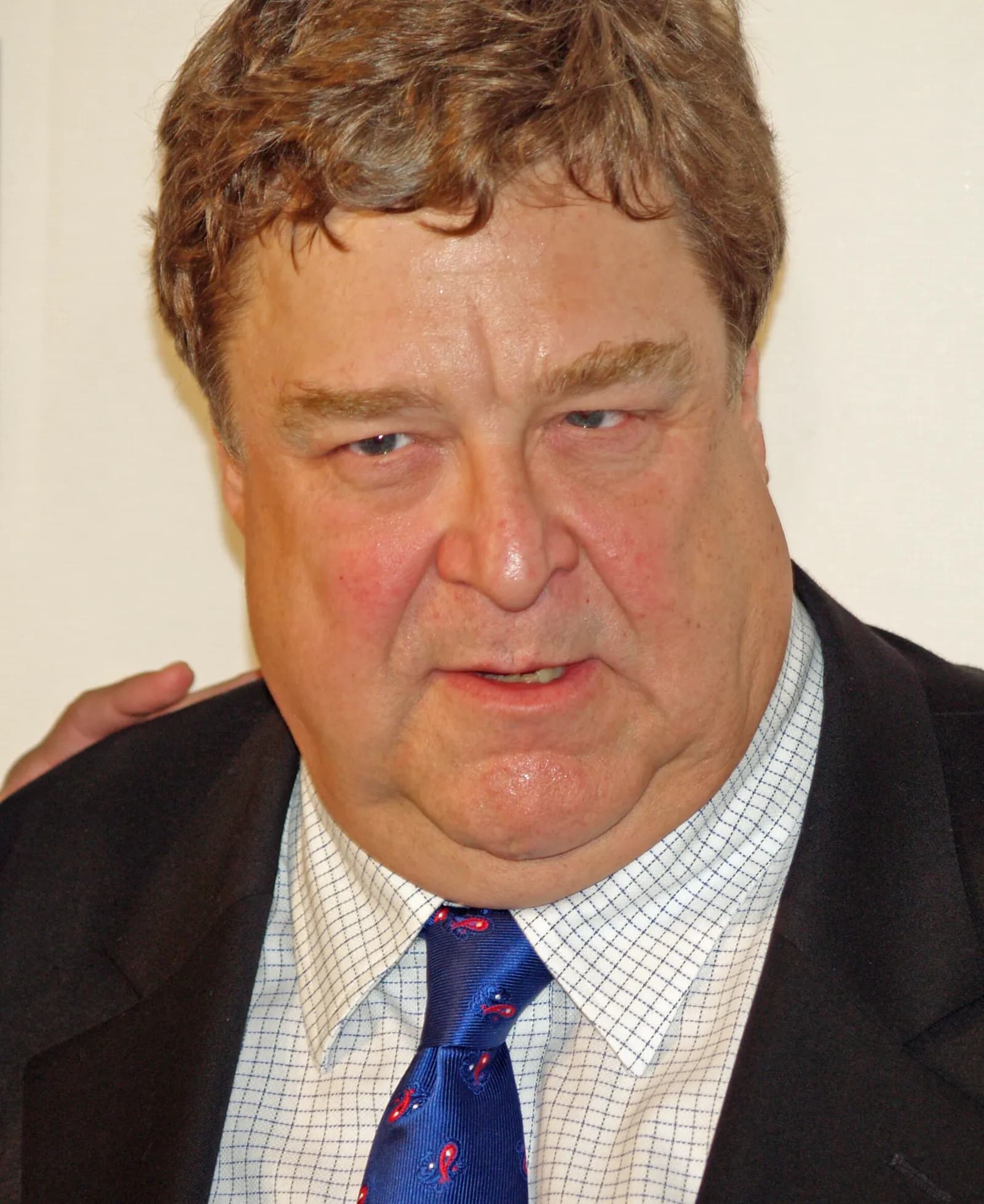 Close-up portrait of John Goodman in a black suit jacket, white checked shirt and blue patterned tie, looking slightly downwards.