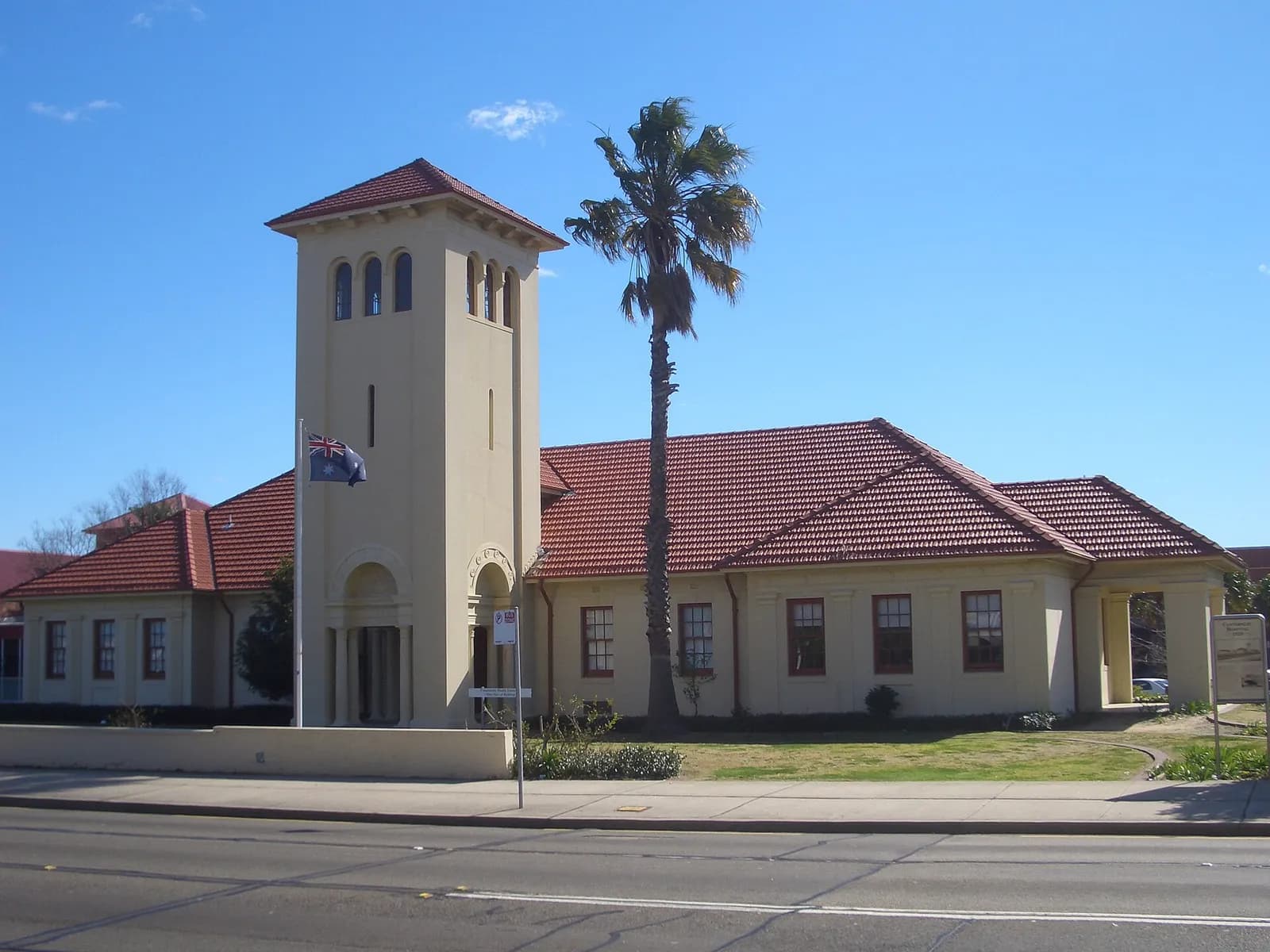 Exterior of a cream-coloured hospital building with a square entry tower, red-tiled pitched roofs, arched windows and doorways, an Australian flag on a pole and a tall palm tree beside the entrance.