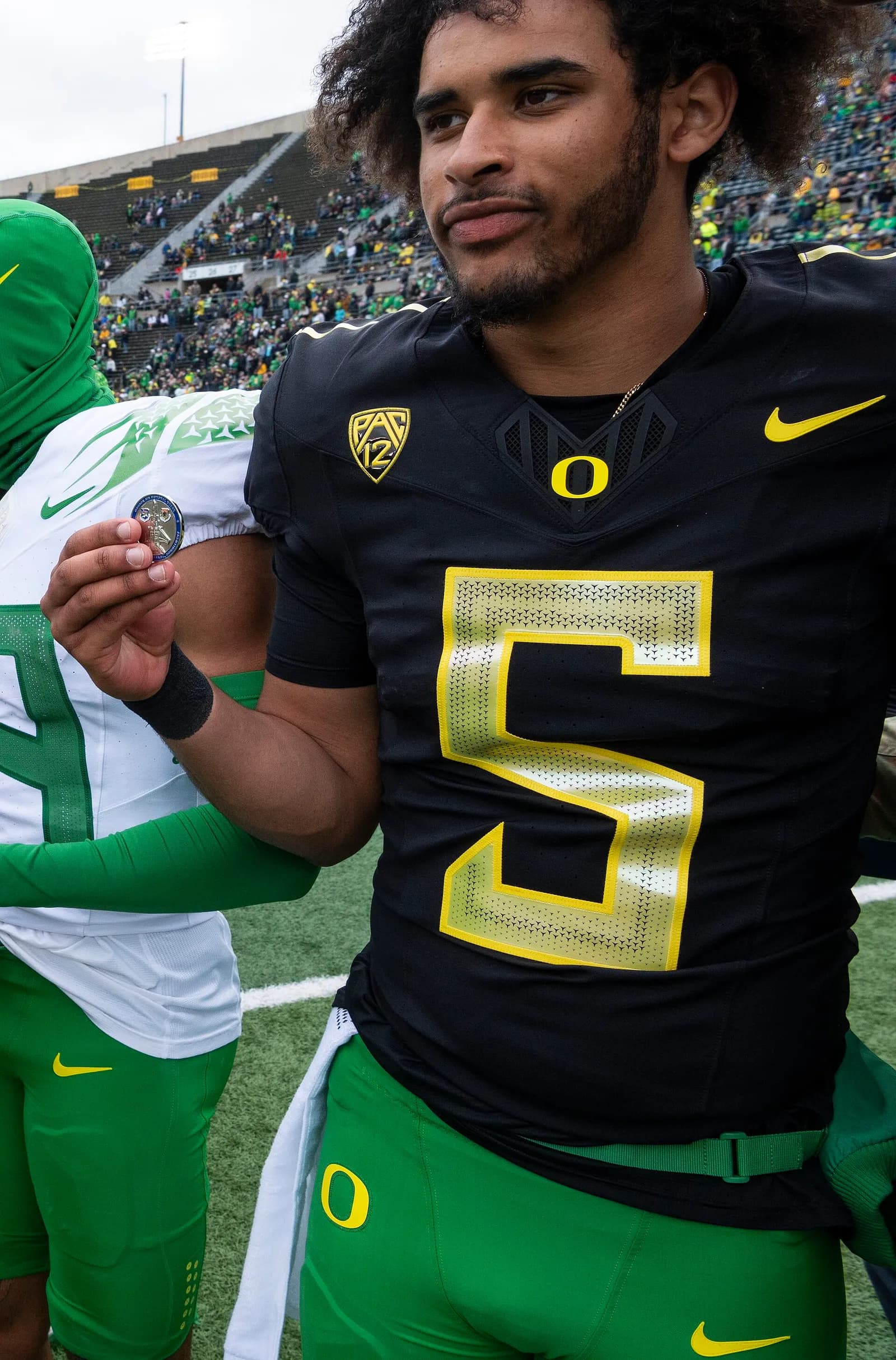 Close-up of a curly-haired American football player in a black Oregon Ducks jersey with a large yellow number 5 and a Pac-12 patch, wearing green shorts, with a person in green and white partly visible behind him and stadium seating in the background.