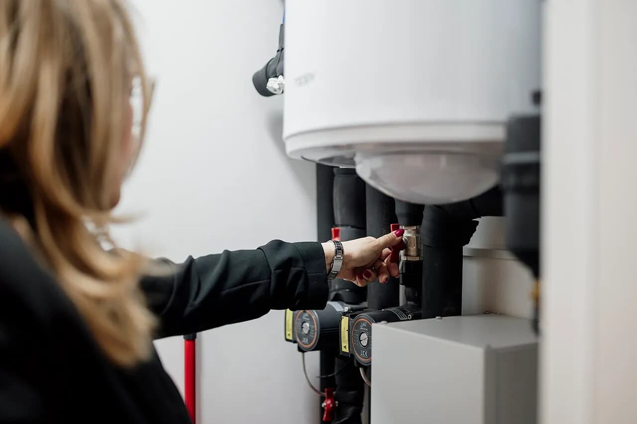 A person adjusts a valve among plumbing equipment in a utility room.