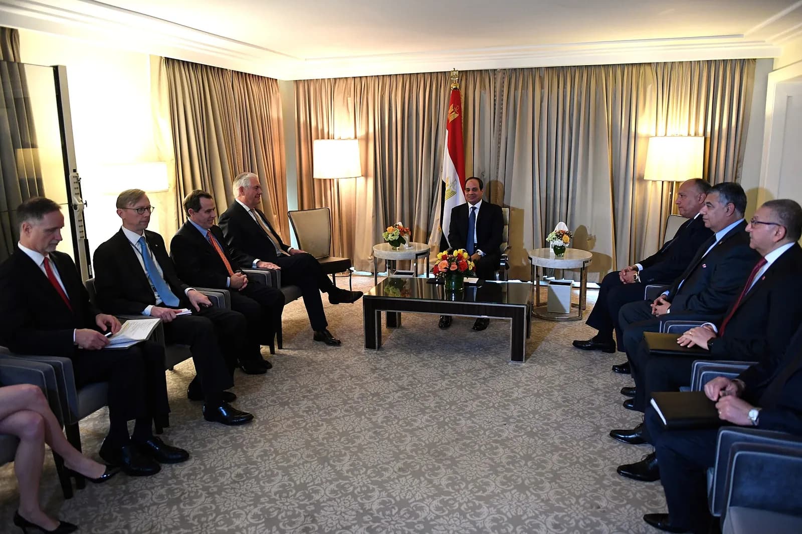 A dark-suited man sits alone behind a glass coffee table beneath an Egyptian flag, facing two rows of seated men in suits.