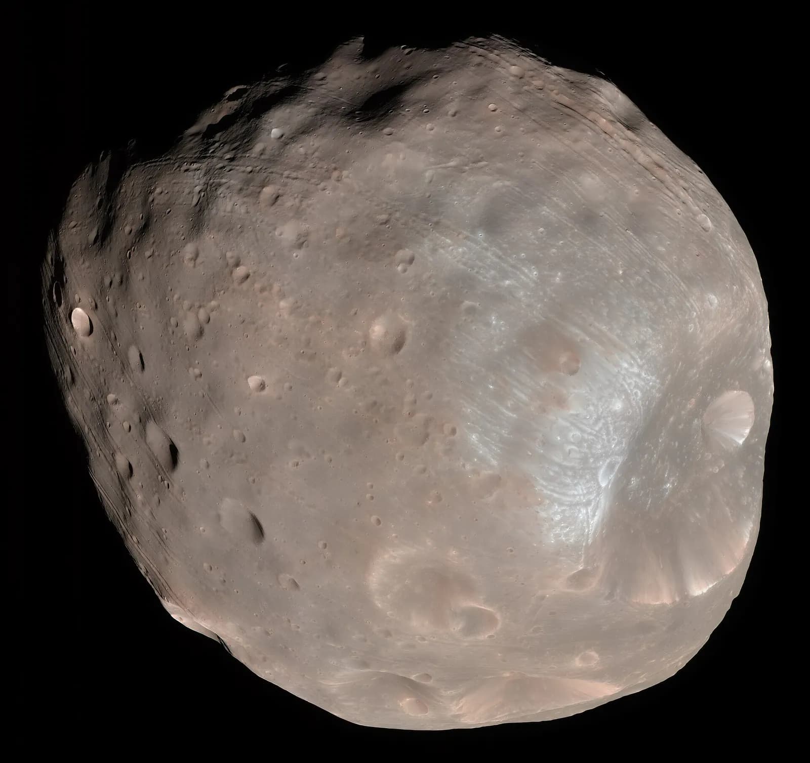 A potato-shaped, cratered grey-brown moon against black space, with long parallel grooves across its surface and shadowed craters on its upper left edge.