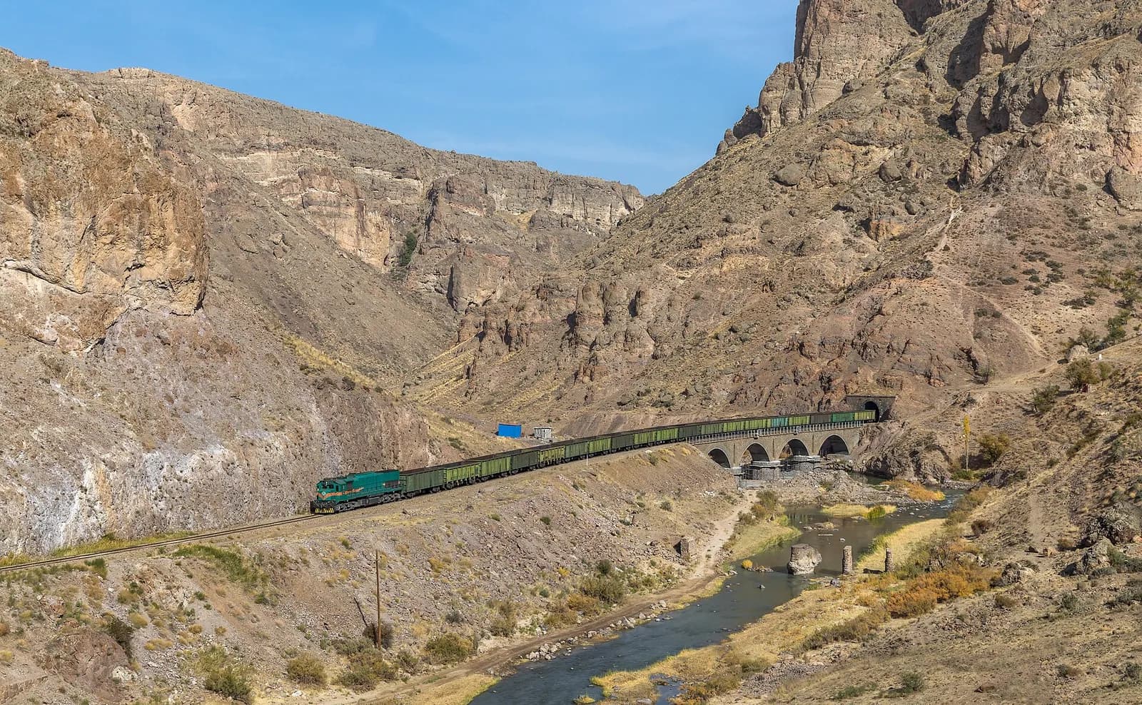 An Iranian Railways locomotive hauling a freight train across a bridge in a river canyon.