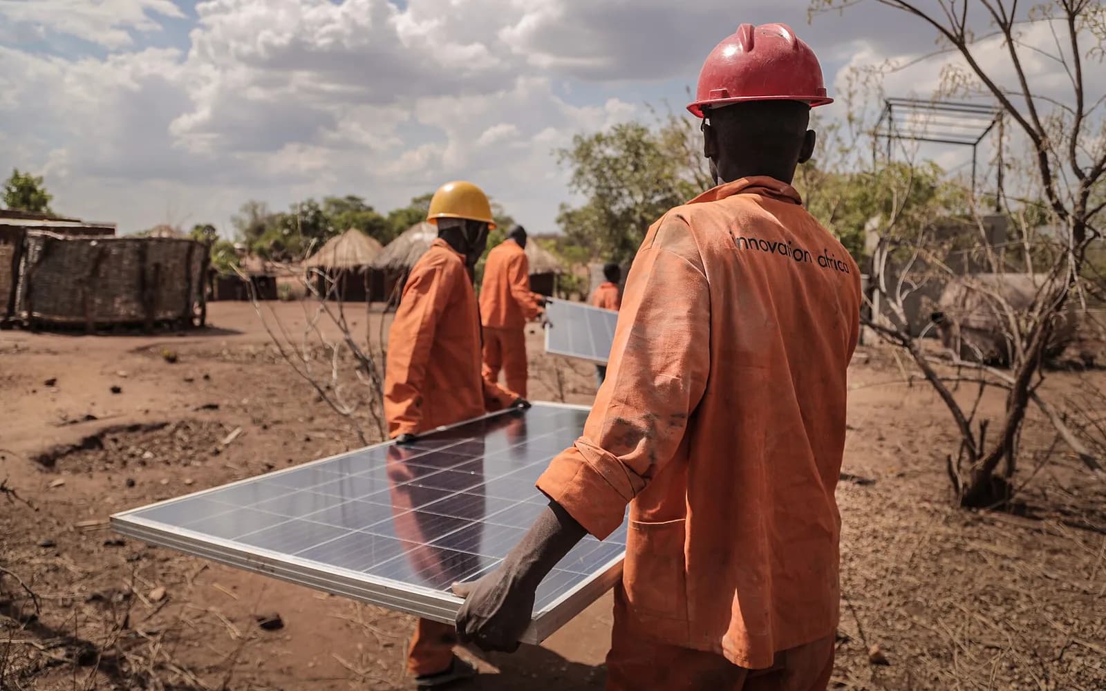 Workers in orange workwear carry solar panels across a rural site.